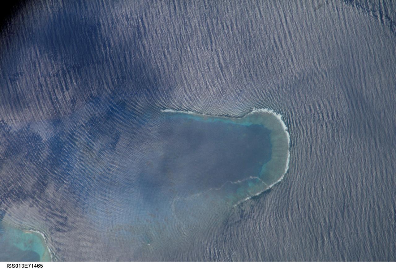 ISS013-E-71465 (27 Aug. 2006) --- Bajo Nuevo Reef in the western Caribbean Sea is featured in this image photographed by an Expedition 13 crewmember on the International Space Station. The Bajo Nuevo reef is approximately 26 kilometers long, comprising two separate U-shaped cays, with only tiny sandy islands associated with them. The eastern cay, shown in this image, encloses a lagoon. Protected from open-sea swells, the water surface is calmer and some back reef detail is visible within the lagoon. Cloud shadows darken the top left corner. The clearest feature is the broad pattern of swells which show two slightly different orientations, producing a tightly crossed pattern. These swells are oriented transverse to the wind direction and were probably set in motion by two different storms hundreds of kilometers east of the reef.  A single irregular line of light color--also suggestive of surface oil--traces a path along the south side of the lagoon.