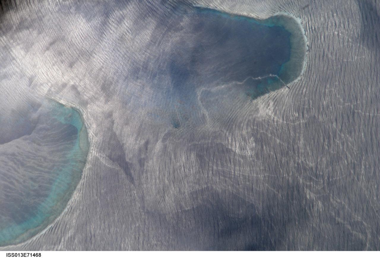 ISS013-E-71468 (27 Aug. 2006) --- Bajo Nuevo Reef in the western Caribbean Sea is featured in this image photographed by an Expedition 13 crewmember on the International Space Station. The Bajo Nuevo reef is approximately 26 kilometers long, comprising two separate U-shaped cays, with only tiny sandy islands associated with them. This detailed view reveals small, low islands that fringe the upwind side (image right) of the lagoon. These islands seem dwarfed by wave swells, which almost appear to wash over them. The clearest feature is the broad pattern of swells which show two slightly different orientations, producing a tightly crossed pattern. These swells are oriented transverse to the wind direction and were probably set in motion by two different storms hundreds of kilometers east of the reef. A single irregular line of light color--also suggestive of surface oil--traces a path along the south side of the lagoon.