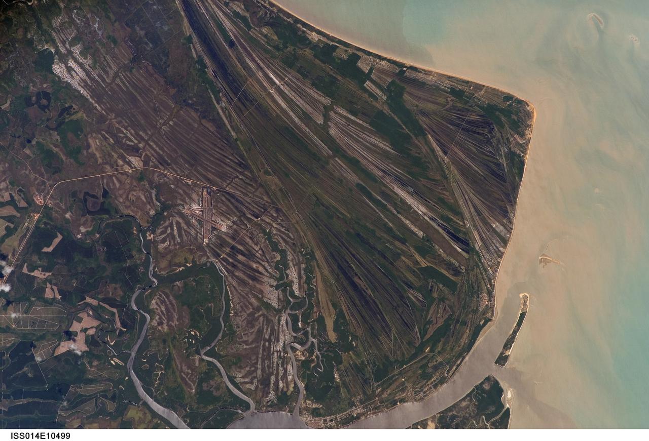 ISS014-E-10499 (23 Dec. 2006) --- Caravelas strandplain, Bahia Province, Brazil is featured in this image photographed by an Expedition 14 crewmember on the International Space Station. This view highlights a flat coastal landform known as a strandplain, or ancient shoreline. The image is dominated by numerous, fine parallel lines (trending diagonally from upper left to lower right), each of which is an ancient shoreline made up of sand transported from rivers to the north. The strandplain has been generated by these narrow shorelines accumulating against one another, line by line in their dozens or even hundreds, over thousands of years.  The shorelines can be grouped into at least four packets depending on the crosscutting relationships - younger packets will cut into or stratigraphically overlay older packets. These relationships indicate that the youngest packet lies nearest the coast (furthest right) and the oldest packet lies north of the city of Caravelas (bottom). The Caravelas River flows into the Atlantic Ocean at the bottom of the view. Sediment from this river, and from the current shoreline, produces the light browns and dun colors visible offshore. On the day this image was taken the river water was relatively clear; clear water (gray) is visible flowing out of the main river mouth, and also off to one side to the south over a levee. The Caravelas airport appears near the middle of the view, and is built on one of the ancient shoreline packets. Caravelas itself, a fishing town of about 20,000 inhabitants, lies on an estuary and was once a flourishing whaling center--the prominent cape at top right is known as Ponta da Baleia (Whale Point).