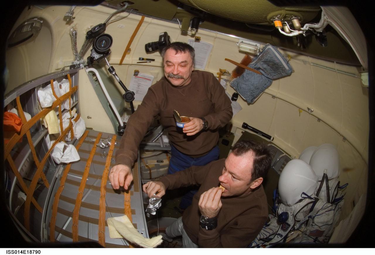 ISS014-E-18790 (29 March 2007) --- Astronaut Michael E. Lopez-Alegria (foreground), Expedition 14 commander and NASA space station science officer; cosmonaut Mikhail Tyurin, Soyuz commander and flight engineer representing Russia's Federal Space Agency; and astronaut Sunita L. Williams (out of frame), flight engineer, share a meal in the orbital module of the Soyuz 13 (TMA-9) spacecraft docked to the International Space Station. Later the crewmembers donned their Russian Sokol launch and entry suits and relocated the Soyuz from the Zarya Module nadir port to the Zvezda Service Module aft port in preparation for the arrival of Expedition 15.