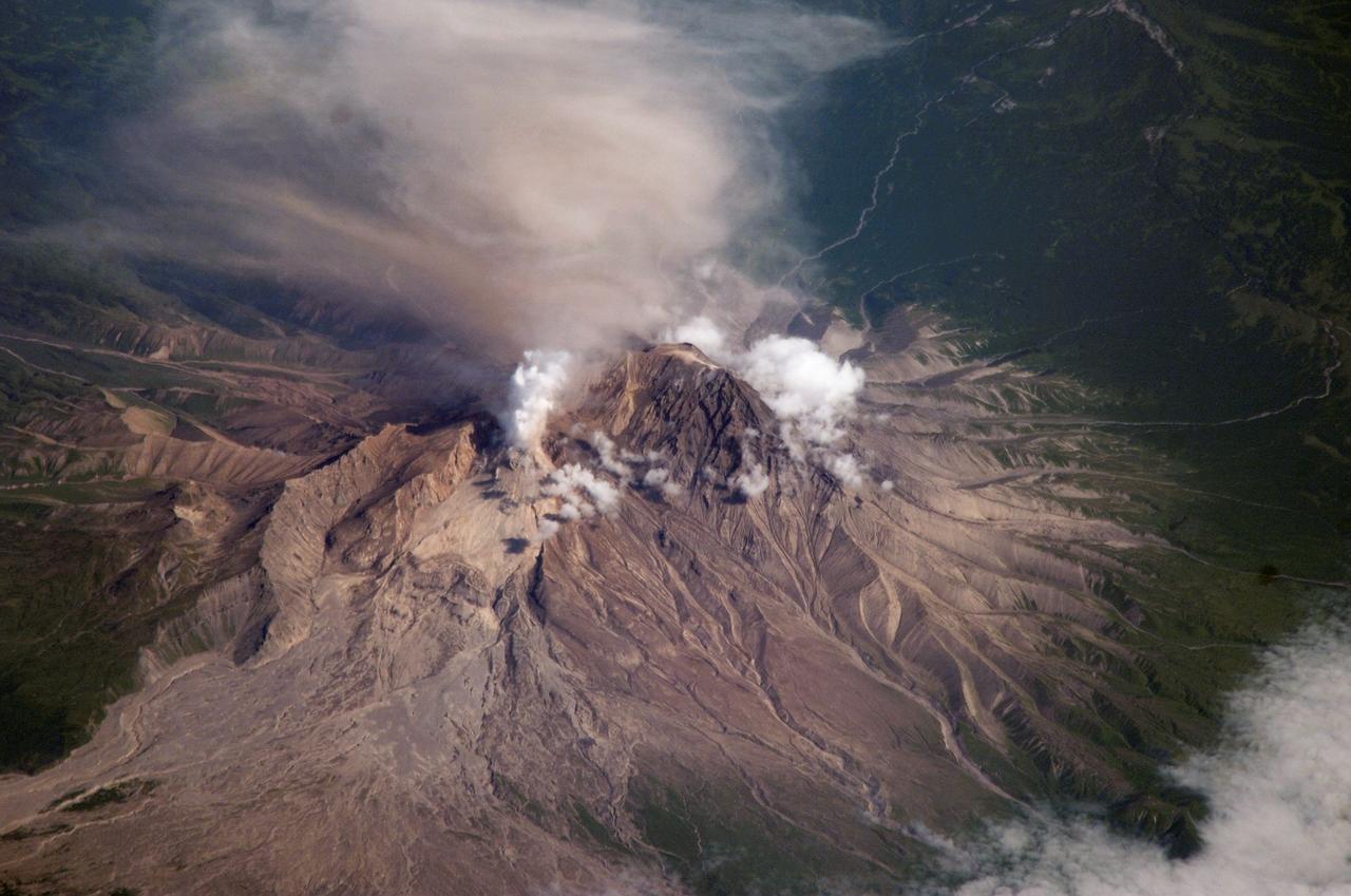 ISS015-E-16913 (10 July 2007) --- Shiveluch Volcano, Kamchatka, Russian Far East is featured in this image photographed by an Expedition 15 crewmember on the International Space Station. Shiveluch is one of the biggest and most active of a line of volcanoes along the spine of the Kamchatka peninsula in easternmost Russia. In turn the volcanoes and peninsula are part of the tectonically active "Ring of Fire" that almost surrounds the Pacific Ocean, denoted by active volcanoes and frequent earthquakes. Shiveluch occupies the point where the northeast-trending Kamchatka volcanic line intersects the northwest-trending Aleutian volcanic line. Junctions such as this are typically points of intense volcanic activity. According to scientists, the summit rocks of Shiveluch have been dated at approximately 65,000 years old.  Lava layers on the sides of the volcano reveal at least 60 major eruptions in the last 10,000 years, making it the most active volcano in the 2,200 kilometer distance that includes the Kamchatka peninsula and the Kuril island chain. Shiveluch rises from almost sea level to well above 3,200 miles (summit altitude 3,283 miles) and is often capped with snow. In this summer image however, the full volcano is visible, actively erupting ash and steam in late June or early July, 2007. The dull brown plume extending from the north of the volcano summit is most likely a combination of ash and steam (top). The two larger white plumes near the summit are dominantly steam, a common adjunct to eruptions, as rain and melted snow percolate down to the hot interior of the volcano. The sides of the volcano show many eroded stream channels. The south slope also reveals a long sloping apron of collapsed material, or pyroclastic flows. Such debris flows have repeatedly slid down and covered the south side of the volcano during major eruptions when the summit lava domes explode and collapse (this occurred during major eruptions in 1854 and 1964). Regrowth of the forest on the south slope (note the contrast with the eastern slope) has been foiled by the combined effects of continued volcanic activity, instability of the debris flows and the short growing season.