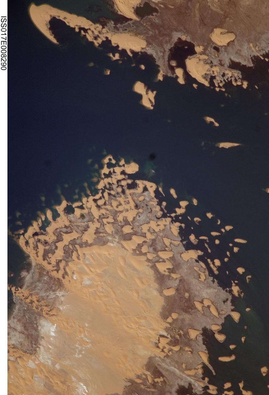 ISS017-E-008290 (31 May 2008) --- Toshka Lakes in southern Egypt are featured in this image photographed by an Expedition 17 crewmember on the International Space Station. In the late 1990s, Egypt's new manmade Toshka Lakes, fed from Lake Nasser via a new canal, grew and spilled into new basins to become four major and two smaller waterbodies extending 120 kilometers across the desert west of the Nile River in southern Egypt. Starting in 2002, astronauts have seen the lakes slowly decline, with the telltale ring of darker, moistened ground showing the previous higher water levels. The rise and fall of Toshka Lakes, and the economic development surrounding the region, are dependent on climate fluctuations and water agreements with upstream countries that, in turn, determine the long-term water flow in the lower Nile. This view, covering a distance of 11.3 kilometers from bottom to top, shows shoreline detail of the third large lake. Lake water appears deep blue to blue-green, and parallel with the shoreline is a wide brown zone which was under water until 2002. Most of the bright yellow sand dunes in the view are re-emerging as the lake level drops, most outlined visibly by thin wet margins. Still-submerged dunes can be seen as small gray blebs offshore (left center). The summit of the biggest horn-shaped dune (top left) was an island more than 5 kilometers off shore in images from 2001. The dune pattern gives a strong sense of dune migration southwards, from top left towards lower right (horns of crescent dunes point in the direction of dune movement). Dominant northerly winds drive the dunes southward, except, of course, when they are under water.
