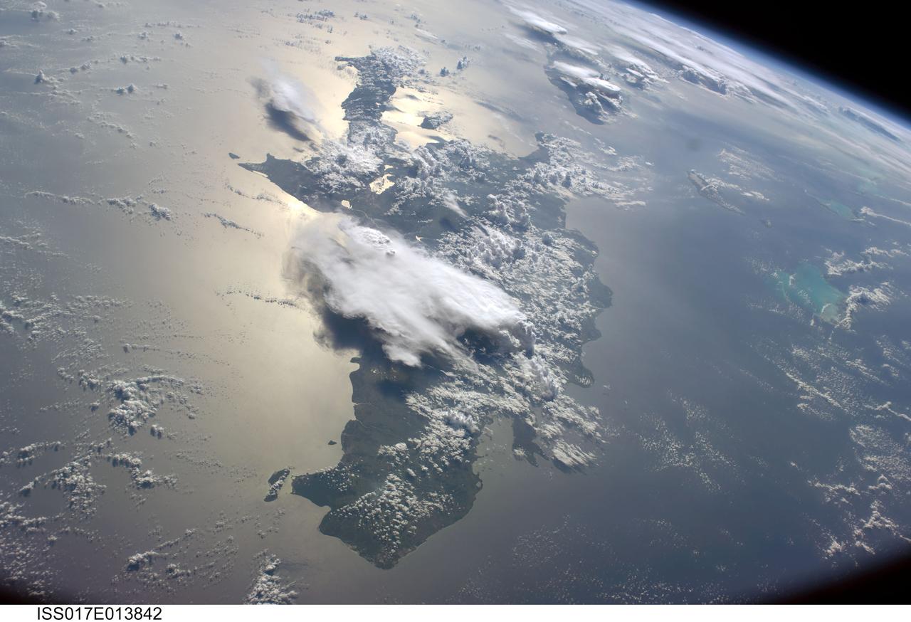 ISS017-E-013842  (19 Aug. 2008) ---   A serene view of a portion of the Greater Antilles islands in the Caribbean Sea.  The island of Hispaniola is in the foreground, comprised of the Dominican Republic and Haiti (in the sunglint).  Looking toward the horizon, the eastern tip of Cuba is seen through the clouds and thunderstorms that are scattered over the islands.  The light blue shallower areas, to the right or north of Hispaniola and extending toward the Earths limb, are the Turks and Caicos islands and the Acklins Islands.  The Great Inagua island is off the coast of Haiti.  These islands are located along the geological border of the North American Plate (to the right) and the Caribbean Plate (to the left). The photo was taken by the Expedition 17 crew onboard the International Space Station on Aug 19, 2008 with a 28 mm lens.