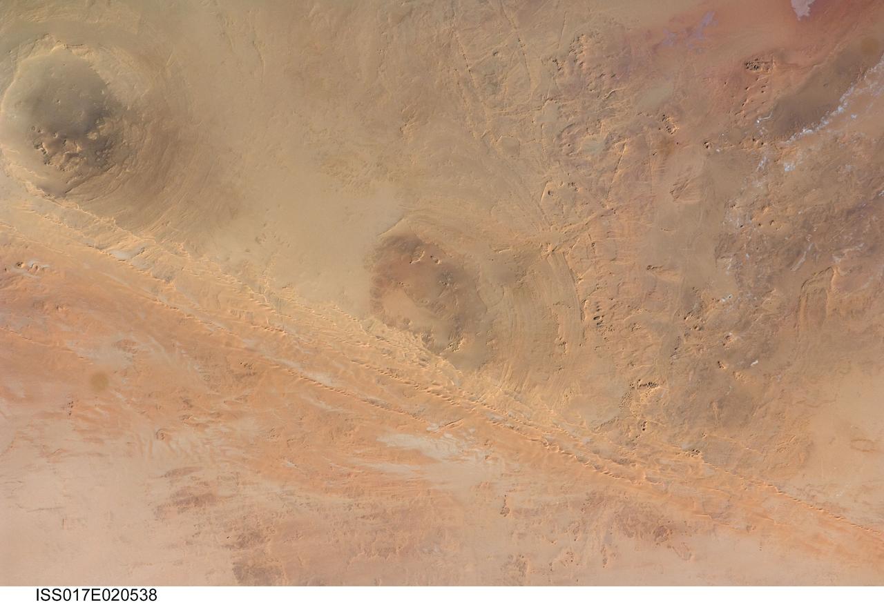ISS017-E-020538 (21 Oct. 2008) --- Arkenu Craters 1 and 2 in Libya are featured in this image photographed by an Expedition 17 crewmember on the International Space Station. Geologists often study features on Earth, such as impact craters, to gain insight into processes that occur on other planets. On Earth, more than 150 impact craters have been identified on the continents, but only a few of these are classified as double impact craters. One such example, the Arkenu Craters in northern Africa, is shown in this image. Arkenu 1 and 2 are double impact structures located in eastern Libya (22.04 degrees north latitude and 23.45 degrees east longitude) in the Sahara desert, with diameters of approximately 6.8 kilometers and 10.3 kilometers, respectively. The craters are unusual in that they both exhibit concentric annular ridge structures (gray circles in the image indicate the position of the outermost visible ridges). In many terrestrial complex craters these features are highly eroded and no longer visible. While the circular structure of these features had been noted, the impact origin hypothesis was strengthened in December 2003 when a field team observed shatter cones -- conical-shaped features in rocks created by the high shock pressures generated during impact. Large outcrops of impact breccias -- a jumble of rock fragments generated at the impact site that are now cemented together into an identifiable rock layer -- were also observed by the field team. Two impactors, each approximately 500 meters in diameter, are thought to have created the craters. According to scientists, the age of the impact event has been dated as occurring less than 140 million years ago. While the presence of shatter cones and impact breccias is generally considered to be strong evidence for meteor impact, some scientists now question the interpretation of these features observed at the Arkenu structures and suggest that they were caused by erosive and volcanic processes. At present, both craters are being crossed by linear dunes extending northeast-southwest -- the superposition of the dunes across the annular ridges indicates that they are much younger than the craters.