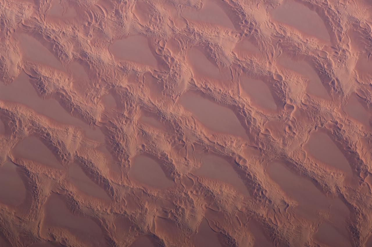 ISS018-E-014770 (20 Dec. 2008) --- Sand dunes in the Marzuq Sand Sea, southwest Libya are featured in this image photographed by an Expedition 18 crewmember on the International Space Station. This detailed view, taken from low Earth orbit, shows classic large and small sand masses of the Central Sahara where wind is a more powerful land-forming agent than water. ?Draa? dunes (from the Arabic for ?arm?) are very large masses of sand and appear here as the broad network of yellow-orange sand masses (the image covers a region approximately 9.4 kilometers wide), with smooth-floored, almost sand-free basins between them. These sand masses lie in the western part of Libya?s vast Marzuq Sand Sea (greater than 60,000 square kilometers, centered at 24.5N 12W). Geologists think that the draa of the Marzuq have probably been formed by winds different from the dominant north/northeast winds of today. Numerous smaller dunes can be seen developed on the backs of the draa. Three distinct dune types can be identified: longitudinal dunes (formed essentially parallel with formative winds from the north); transverse dunes, usually more curved, formed at right angles to the formative wind; and star dunes, in which several linear arms converge towards a single peak. The upwind side of the sand masses appears smoother than the more rippled downwind side. Wind is moving sand grains almost all the time. This means that the draa and the dunes are all moving -- as sand is added on the upwind side and blown off the downwind side. It is well known that small sand masses move much faster than large sand masses. This means that the draa are almost stationary, but that the smaller dunes are moving relatively quickly across their backs. When the dunes reach the downwind side of the draa they are obliterated, their sand being blown across the basins as individual grains.