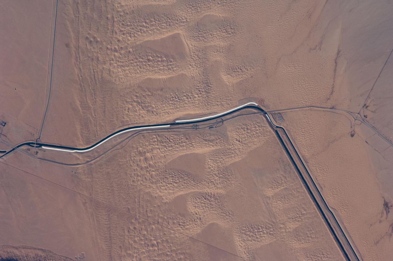 ISS018-E-024949 (31 Jan. 2009) --- The All-American Canal, the largest irrigation canal in the world and a key landmark along the California-Mexico border, is featured in this image photographed by an Expedition 18 crewmember on the International Space Station. This image captures about 15 kilometers of the important infrastructure corridor just west of Yuma, AZ. The prominent dark line crossing the image is the Canal which is crossed, in this view, by Interstate Highway 8. The canal carries 740.6 cubic meters (26,155 cubic feet) of water per second from the Colorado River westward to support the intensive agriculture of California?s Imperial Valley to the northwest, and to nine cities including San Diego, CA. The canal system is the Imperial Valley?s only source of water, and allows irrigation of more than 2,000 square kilometers (500,000 acres) of agricultural fields. The Coachella Canal, one of four main branch canals, leads water north to Imperial Valley. This section of the canal requires constant maintenance. Approximately 68,000 acre-feet of water per year are lost by seepage from the All-American Canal - especially where the canal crosses the great Algodones Dune Field, a portion of which is visible extending from top to bottom in the center of the image. Additionally, dune sand is constantly blown to the southeast, and into the canal. As part of California?s Colorado River Water Use Plan, 37 kilometers (23 miles) of the canal is being lined to prevent water loss by seepage. A recently opened sector parallels the old canal (right) and new lined sectors are under construction (bright lines, center). Engineers have sited new sections of the canal to avoid the worst areas of dune-sand invasion, so that the new configuration will be significantly cheaper to maintain and operate. A new road crosses the dunes and marks the US?Mexico border as part of border fence construction efforts. The margin of the Colorado River floodplain in Mexico is just visible two kilometers south of the border (lower left corner). This floodplain is Mexico?s equivalent of the Imperial Valley in terms of its enormous irrigated agricultural production.