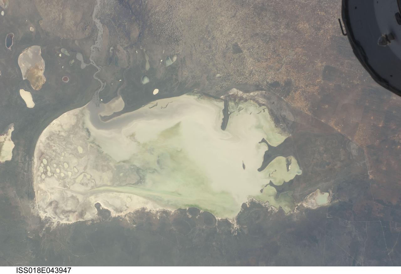 ISS018-E-043947 (26 March 2009) --- Etosha Pan in Namibia is featured in this image photographed by an Expedition 18 crewmember on the International Space Station. Etosha Pan is a large dry lake about 130 kilometers long that dominates Namibia?s Etosha National Park (the sharp edge of the park fence can be seen at right). Small related dry lake beds appear as bright shapes top left, a portion of the International Space Station appears at top right. This view shows the pan as it appeared almost ten years ago. The pan is the low point in a major inland basin in northern Namibia. During occasional flood events, such those experienced in the last nine months, rivers from Angola (the Namibia?Angola boundary lies just outside the top of the image) deliver large quantities of water to the pan. In this image flood water in the Oshigambo River, resulting from recent heavy rains in Angola, appears as a gray stream entering the northwest corner of the pan (top left). The floodwater becomes a thin sheet on the vast salt flat of the pan floor. Algae blooms in the warm water have produced a light-green tinge. Another image of the Oshigambo River mouth in flood can be seen here. Reports on the ground, augmented with orbital unmanned satellite imagery acquired subsequent to this photograph, show that the plains north of the pan are now flooded, causing damage to homesteads, crops and roads. More than 340,000 people have been affected in northern Namibia and approximately 250,000 in southern Angola.