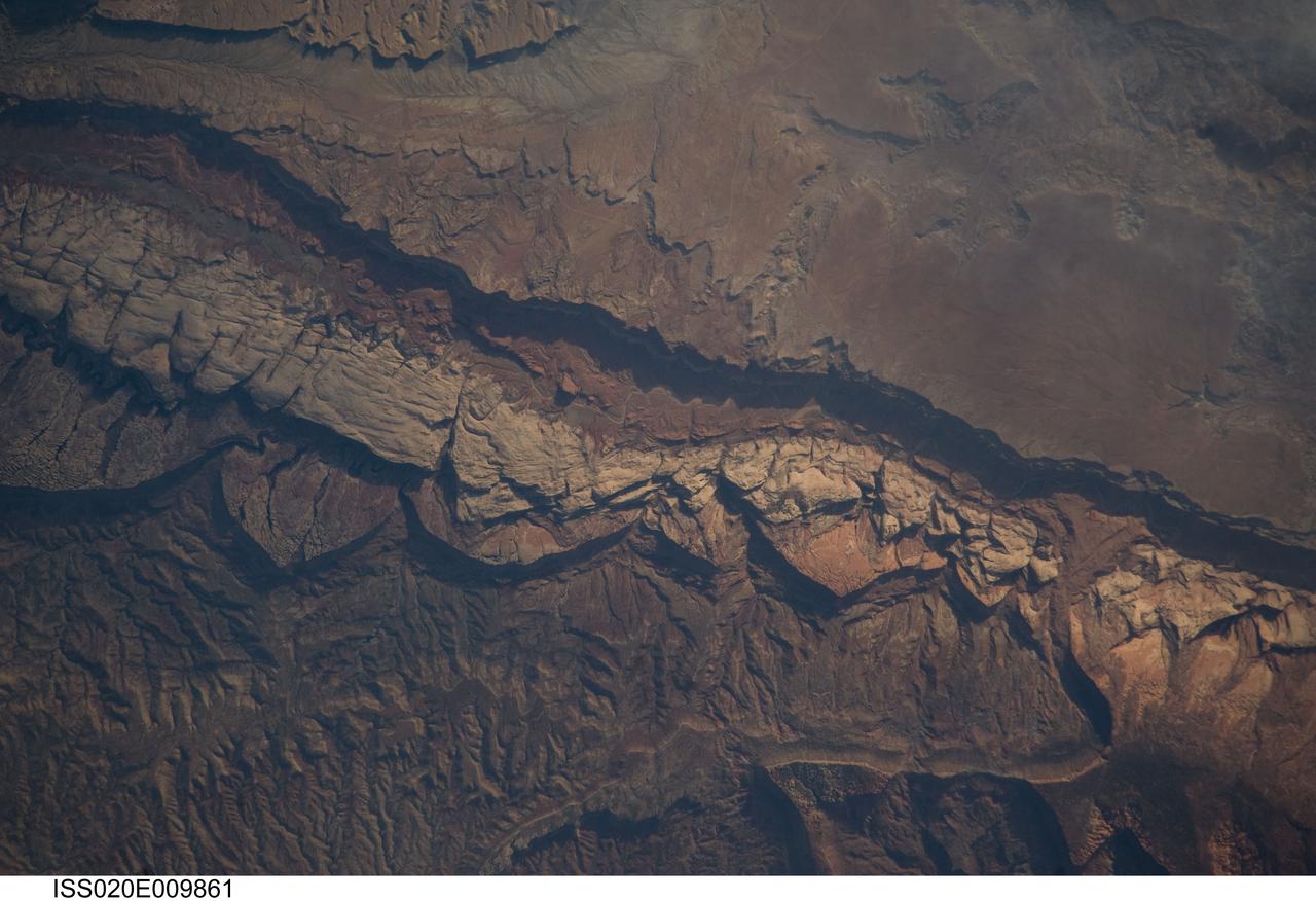 ISS020-E-009861 (14 June 2009) --- Big Thompson Mesa in the Capitol Reef National Park, Utah is featured in this image photographed by an Expedition 20 crew member on the International Space Station. This detailed photograph depicts a portion of Big Thompson Mesa located near the southern end of Capitol Reef National Park. Capitol Reef National Park is located on the Colorado Plateau, a physiographic and geologic province that comprises the adjacent quarters of Arizona, Colorado, New Mexico and Utah. Big Thompson Mesa (upper right) is part of a large feature known as the Waterpocket Fold. The Fold is a geologic structure called a monocline, characterized by generally flat-laying sedimentary rock layers with a steep and highly localized flexure– much like a carpet runner draped over a stair step. Monoclines on the Colorado Plateau are thought to be the result of faulting of stratigraphically lower and more brittle crystalline rocks; while the crystalline rocks were broken into raised or lowered blocks, the overlaying, less brittle sedimentary rocks were flexed without breaking. According to scientists, the portion of the Waterpocket Fold illustrated in this image includes layered rocks formed during the Mesozoic Era (approximately 250 – 65 million years ago) – the oldest layers are at the bottom of the sequence (and also, in this view, the image), with each successive layer younger than the preceding one going upwards in the sequence. Not all of the units present are clearly visible, but some of the major units can be easily distinguished. The bottom half of the image includes the oldest rocks in the view: dark brown and dark green Moenkopi (Trm) and Chinle (Trc) Formations. At center, two strikingly colored units are visible – light red to orange Wingate Sandstone (Jw) and white Navajo Sandstone (Jn). A topographic bench above these units includes reddish brown to brown Carmel Formation (Jc) and Entrada Sandstone (Je). The top of the cliff face above this bench - Big Thompson Mesa - is comprised of brown Dakota Sandstone (Kd). Scientists believe this sequence represents more than 100 million years of deposition. Much younger Quaternary (2.0 million to approximately 10,000 years old) deposits are also present in the view. A regional view of Capitol Reef National Park and the Waterpocket Fold is available here. The area shown in this view is located approximately 65 kilometers to the southeast of Fruita, UT near the southern end of Capitol Reef National Park.