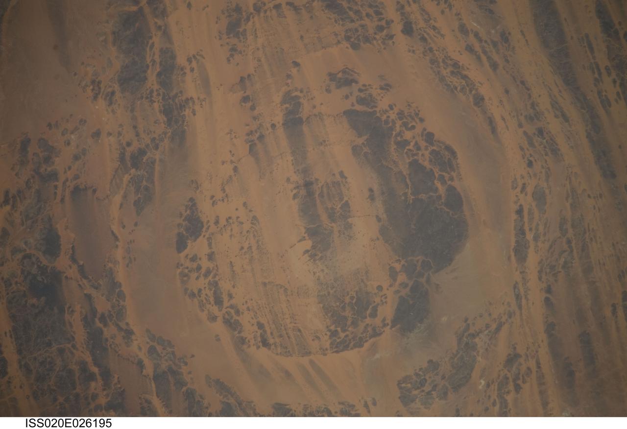 ISS020-E-026195 (25 July 2009) --- Aorounga Impact Crater is featured in this image photographed by an Expedition 20 crew member on the International Space Station. Aorounga Impact Crater is located in the Sahara Desert of north-central Chad and is one of the best preserved impact structures in the world. According to scientists, the crater is thought to be middle or upper Devonian to lower Mississippian (approximately 345 ? 370 million years old) based on the age of the sedimentary rocks deformed by the impact. Spaceborne Imaging Radar (SIR) data collected in 1994 suggests that Aorounga is one of a set of three craters formed by the same impact event. The other two suggested impact structures are buried by sand deposits. The concentric ring structure of the Aorounga crater ? renamed Aorounga South in the multiple-crater interpretation of SIR data ? is clearly visible in this detailed photograph. The central highland, or peak, of the crater is surrounded by a small sand-filled trough; this in turn is surrounded by a larger circular trough. Linear rock ridges alternating with light orange sand deposits cross the image from upper left to lower right; these are called yardangs by geomorphologists. Yardangs form by wind erosion of exposed rock layers in a unidirectional wind field. The wind blows from the northeast at Aorounga, and sand dunes formed between the yardangs are actively migrating to the southwest.