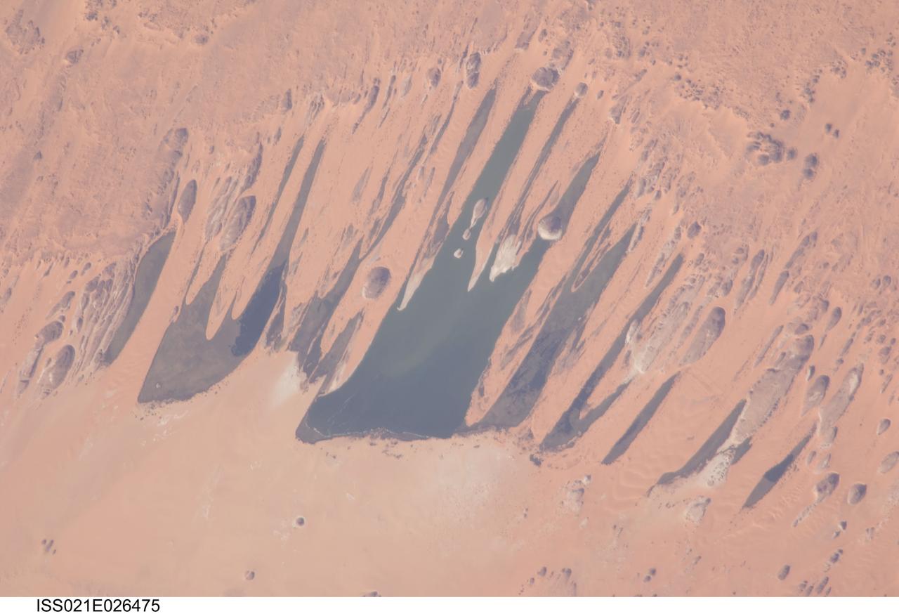 ISS021-E-026475 (14 Nov. 2009) --- Ounianga Lakes in the Sahara Desert, in the nation of Chad are featured in this image photographed by an Expedition 21 crew member on the International Space Station. This view features one of the largest of a series of ten, mostly fresh water lakes in the Ounianga basin in the heart of the Sahara Desert of northeastern Chad. According to scientists, the lakes are the remnant of a single large lake, probably tens of kilometers long that once occupied this remote area approximately 14,800 to 5,500 years ago. As the climate dried out during the subsequent millennia, the lake was reduced in size and large wind-driven sand dunes invaded the original depression dividing it into several smaller basins. The area shown in this image measures approximately 11 x 9 kilometers, with the dark water surfaces of the lake segregated almost completely by orange linear sand dunes that stream into the depression from the northeast. The almost year-round northeast winds and cloudless skies make for very high evaporation (an evaporation rate of greater than six meters per year has been measured in one of the nearby lakes). Despite this, only one of the ten lakes is saline. According to scientists, the reason for the apparent paradox of fresh water lakes in the heart of the desert lies in the fact that fresh water from a very large aquifer reaches the surface in the Ounianga depression in the form of the lakes. The aquifer is large enough to keep supplying the small lakes with water despite the high evaporation rate. Mats of floating reeds also reduce the evaporation in places. The lakes form a hydrological system that is unique in the Sahara Desert. Scientists believe the aquifer was charged with fresh water, and the large original lake evolved, during the so-called African Humid Period (approximately 14,800 to 5,500 years ago) when the West African summer monsoon was stronger than it is today. Associated southerly winds brought Atlantic moisture well north of modern limits, producing sufficient rainfall in the central Sahara to foster an almost complete savanna vegetation cover. Pollen data from lake sediments of the original 50-meters-deep Ounianga Lake suggests to scientists that a mild tropical climate with a wooded grassland savanna existed in the region. This vegetation association is now only encountered 300 kilometers further south. Ferns grew in the stream floodplains which must have been occasionally flooded.  Even shrubs that now occur only on the very high, cool summits (greater than 2,900 meters, greater than 9,500 feet) of the Tibesti Mts. have been found in the Ounianga lake sediments.
