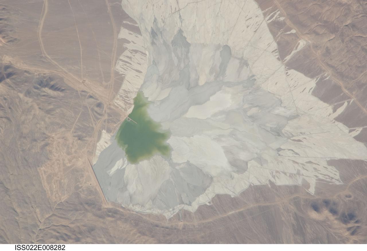 ISS022-E-008282 (9 Dec. 2009) --- One of the world?s leading copper mines, Escondida,  in the Atacama Desert of Chile, is featured in this image photographed by an Expedition 22 crew member on the International Space Station. The copper mining industry is a major part of the Chilean economy. The mine is located 170 kilometers southeast of Chile?s port city of Antofagasta, in the hyper arid northern Atacama Desert at an elevation of 3,050 meters (approximately 10,000 feet) above sea level. Escondida produces mainly copper concentrates; assisted by gravity, the concentrates are piped as slurry down to the smaller port of Coloso just south of Antofagasta where they are dewatered for shipping. The photograph features a large light tan and gray waste or ?spoil? materials impoundment area (center) of the mine complex. The copper-bearing waste, which is a large proportion of the material excavated from open pits to the north (not in frame), is poured into the impoundment area as a liquid (green region at photo?s center), and dries to the lighter-toned spoil seen in the image. The spoil is held behind a retaining dam, just a little more than one kilometer in length, visible as a straight line at lower left. ?Escondida? means ?hidden? in Spanish, and refers to the fact that the copper ore body was buried beneath hundreds of meters of barren rock and had to be located by a laborious drilling program following a geologic trend established from other copper occurrences.