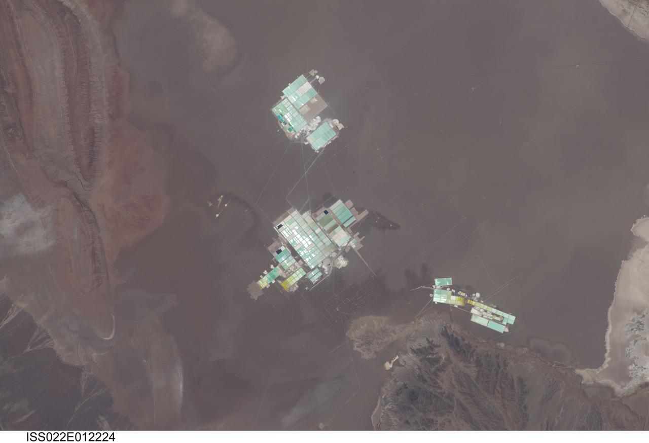 ISS022-E-012224 (15 Dec. 2009) --- Evaporation ponds in the Salar de Atacama, Chile are featured in this image photographed by an Expedition 22 crew member on the International Space Station. The Salar (salt flat) de Atacama in Chile is an enclosed basin with no drainage outlets. While the grey-brown surface of the Salar is flat and desiccated, mineral-rich brines - water with a high percentage of dissolved salts - are located below the surface. The subsurface brines of the Salar de Atacama are particularly rich in lithium salts. Lithium is an essential component of advanced batteries and medicines, among many other uses. The brines are pumped to the surface through a network of wells and into large shallow evaporation ponds ? three such evaporation pond facilities are visible in the center of the image. Color variations in the ponds are due to varying amounts of salts relative to water. The dry and windy climate enhances evaporation of the water, leaving concentrated salts behind for extraction of the lithium. The Salar de Atacama is located in the southern half of the Atacama Desert ? with no historical or current records of rainfall in some parts of this desert, it is considered to be one of the driest places on Earth. This photograph illustrates the central portion of the Salar de Atacama. It is bounded by brown to grey-brown folded and faulted strata of the Cordillera de la Sal to the northwest (upper left) and darker bedrock of the Cordon de Lila to the south (lower right).