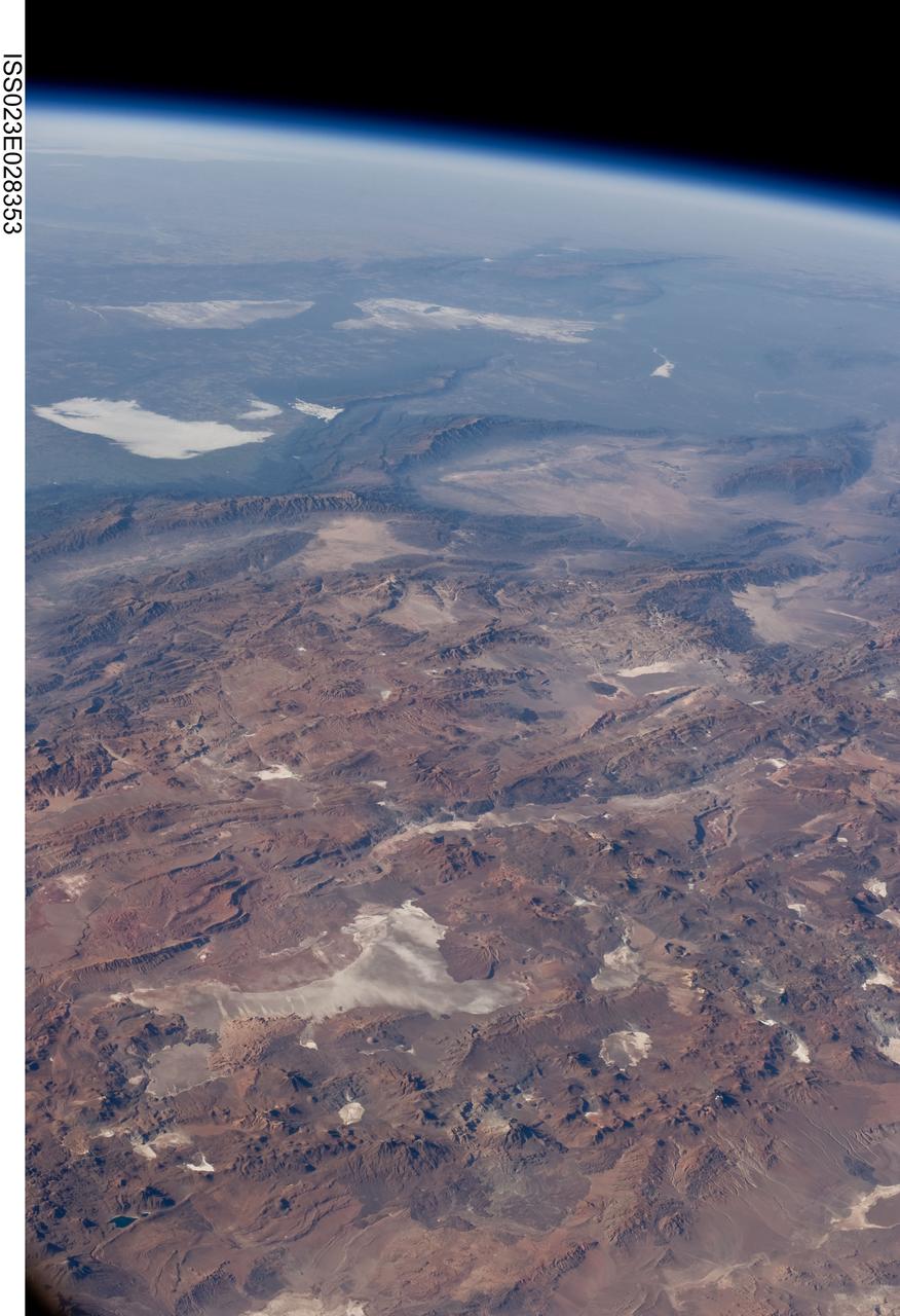 ISS023-E-028353 (26 April 2010) --- Central Andes Mountains, Salar de Arizaro, Argentina are featured in this image photographed by an Expedition 23 crew member on the International Space Station. The high plains (3,000 to greater than 5,000 meters elevation, 13,000 to 19,000 feet) of the Andes Mountains, also known as the Puna, appear in the foreground of this photograph, with a line of young volcanoes facing the much lower Atacama Desert (1,000–2,000 meters elevation). Several large dry lakes, marked by light-toned salt crusts, occupy the basins between major thrust faults in the Puna. Salar de Arizaro (foreground) is the largest of the dry lakes in this view (salar means waterless salt flat in Spanish). This panorama was taken by a station crew member looking southeast across the South American continent when the ISS was almost directly over the Atacama Desert near Chile’s Pacific coast. The Atlantic Ocean coastline (River Plate where Argentina’s capital city of Buenos Aires is located) is dimly visible at top left. A striking geological and landscape contrast is visible at center which separates two distinct geological zones, namely the Puna and the Sierras Pampeanas. The Sierras Pampeanas Mountains are lower in elevation and have few young volcanoes, in contrast to the Puna. Sharp-crested ranges are separated by wide, low valleys in this region. The Salinas Grandes—ephemeral shallow salt lakes at top left (salina means salt lake in Spanish) — occupies one of these valleys. The general color change from reds and browns in the foreground to blues and greens in the upper part of the image reflects the major climatic regions, namely the deserts of the Atacama and Puna, versus the low grassy plains of central Argentina where rainfall is sufficient to promote lush prairie grass growth—known famously as the pampas in Argentina. The Salinas Grandes mark an intermediate semiarid region. What accounts for the changes in landscape? The geology of this part of the Andes is a result of the eastward subduction of the Nazca tectonic plate underneath South America. Investigations using seismic data suggest that the Puna is underlain by a steeply dipping sector of the subducting Nazca plate. The Sierras Pampeanas zone however, is underlain by a sector of the Nazca plate that is almost horizontal, possibly due to the subduction of a submarine mountain range known as the Juan Fernandez Ridge. In the simplest terms, ridges are topographic highs that are difficult to stuff down subduction zones, with profound effects on the volcanism and structures of the upper plate.