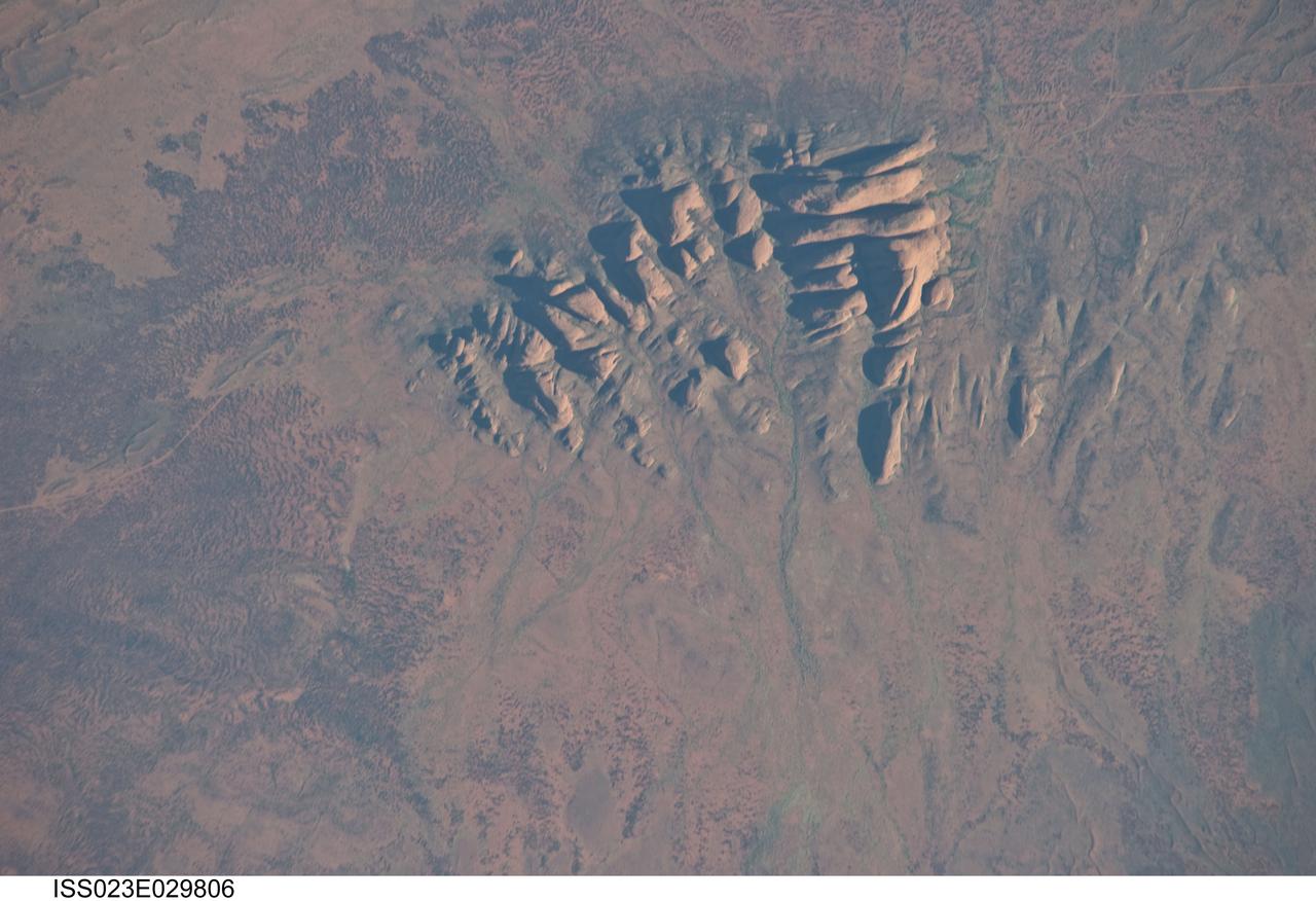 ISS023-E-029806 (30 April 2010) --- Kata Tjuta, Australia is featured in this image photographed by an Expedition 23 crew member on the International Space Station. Located in the Northern Territory of Australia, Uluru – Kata Tjuta National Park hosts some of the world’s most spectacular examples of inselbergs, or isolated mountains. The most famous of these inselbergs is Uluru (also known as Ayers Rock). An equally massive inselberg located approximately 30 kilometers to the northwest is known as Kata Tjuta– like Uluru, this is a sacred site to the native Anangu or Aboriginal people. Explorers named the highest peak Mount Olga, with the entire grouping of rocks informally known as “the Olgas”.  Mount Olga has a peak elevation of 1,069 meters above sea level, making it 206 meters higher than Uluru. Kata Tjuta is comprised of gently dipping Mount Currie Conglomerate, a sedimentary rock that includes abundant rounded fragments of other rock types (here, primarily granite with less abundant basalt and rhyolite) in a coarse sandy matrix. Geologists interpret the Mount Currie Conglomerate as a remnant of a large fan of material rapidly eroded from mountains uplifted approximately 550 million years ago. Subsequent burial under younger sediments consolidated the eroded materials to form the conglomerate exposed at the surface today. In this photograph, afternoon sunlight highlights the rounded summits of Kata Tjuta against the surrounding sandy plains. Sand dunes are visible at upper right; while in other areas (image top and image left) sediments washed from the rocks have been anchored by a variety of grasses and bushes adapted to the arid climate. Green vegetation in the ephemeral stream channels that drain Kata Tjuta (bottom center) provides colorful contrast with the red rocks and surrounding soils. Large gaps in the rocks (highlighted by shadows) are thought to be fractures that have been enlarged due to erosion.
