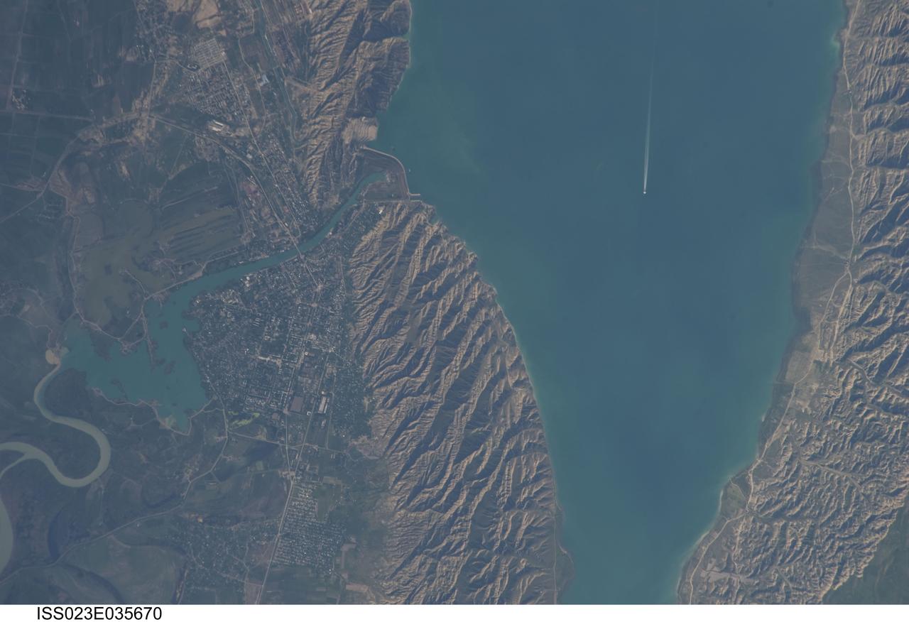 ISS023-E-035670 (8 May 2010) --- Mingachevir Reservoir, Azerbaijan is featured in this image photographed by an Expedition 23 crew member on the International Space Station. This detailed photograph highlights the southern Mingachevir Reservoir located in north-central Azerbaijan. The Mingachevir Reservoir occupies part of the Kura Basin, a topographic depression located between the Greater Caucasus Mountains to the northeast and the Lesser Caucasus Mountains to the southwest. According to scientists, folded layers of relatively young (less than 5.3 million years old) sedimentary rock, explosive volcanic products (ash and tuff), and unconsolidated sediments form the gray hills along the northern and southern shorelines of the reservoir (center and right). Afternoon sun highlights distinctive parallel patterns in the hills that are the result of water and wind erosion of different stratigraphic layers exposed at the surface. The nearby city of Mingachevir (left) is split by the Kur River after it passes through the dam and hydroelectric power station complex at top center. The current city was built in support of the hydroelectric power station constructed as part of the then Soviet Union?s energy infrastructure for the region. Today, Mingachevir is the fourth largest city in Azerbaijan (by population) and has become a cultural and economic center of the country. The reservoir held approximately 15 billion cubic meters of water at the time this image was taken, with a total engineered capacity of 16 billion cubic meters. The width of the reservoir illustrated here is approximately 8 kilometers; a jet flying over the reservoir and its contrail are visible midway between the opposing shorelines.