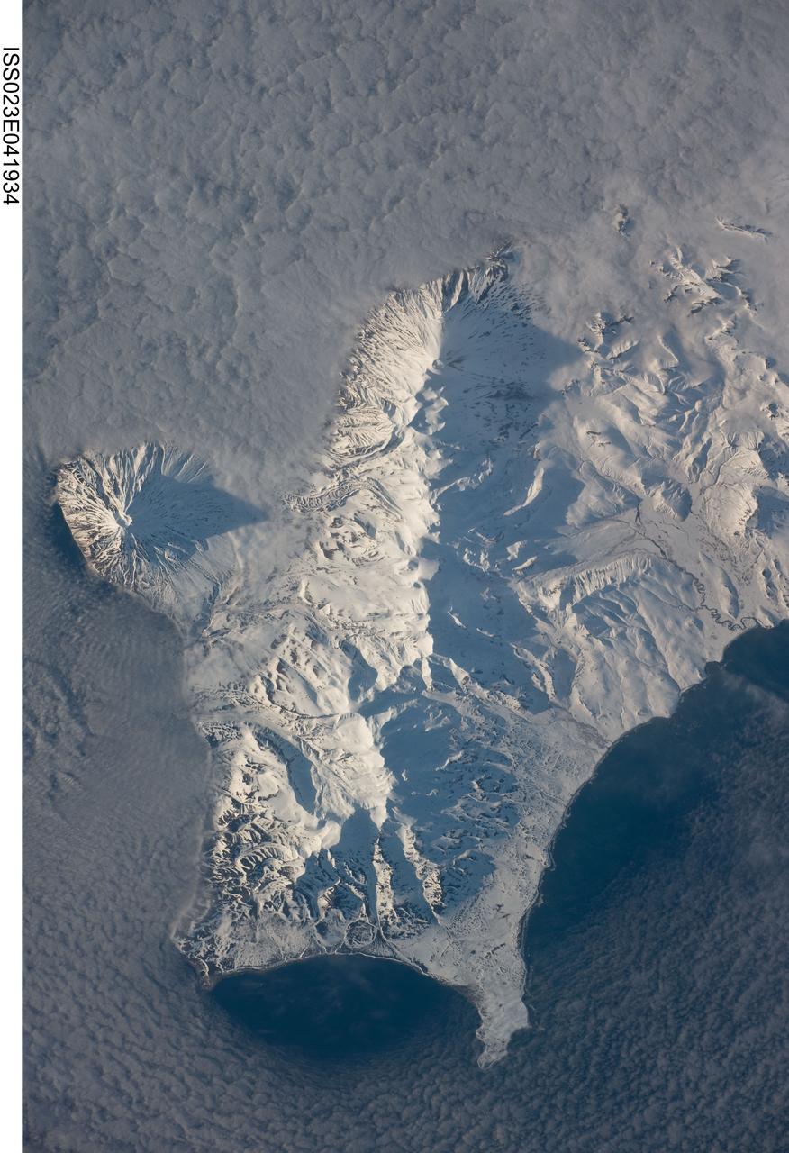 ISS023-E-041934 (12 May 2010) --- Southern Paramushir Island in the Kuril island chain in Russia is featured in this image photographed by an Expedition 23 crew member on the International Space Station. The Kuril island chain consists of a line of volcanoes, many of which have been historically active, that extends from the Kamchatka Peninsula to northern Japan. This line of island volcanoes is known to geologists as an island arc. Island arcs form along an active geologic boundary, typically marked by a deep undersea trench, between two tectonic plates with one being driven beneath the other (a process called subduction). Magma generated by this process feeds volcanoes ? and eventually, volcanic islands ? over the subduction boundary. Paramushir Island in the northern Kurils is an example of a large island built by several volcanoes over geologic time. This photograph shows the southern end of Paramushir Island after a snowfall. There are four major volcanic centers that form this part of the island. Fuss Peak (center left) is an isolated stratovolcano connected to the main island via an isthmus. The last recorded historical eruption of Fuss Peak was in 1854. The southern tip of the island is occupied by the Karpinsky Group of three volcanic centers.  A minor eruption of ash following an earthquake occurred on this part of the island in 1952. The Lomonosov Group to the northeast (center) includes four cinder cones and a lava dome that produced several lava flows extending from the central ridge to the east, northeast, west, and southeast. There have been no recorded historical eruptions from the Lomonosov Group of volcanoes. The most recent volcanic activity (in 2008) occurred at the Chikurachki cone located along the northern coastline of the island at top center. The summit of this volcano (1816 meters above sea level) is the highest on Paramushir Island. Much of the Sea of Okhotsk visible in the image is covered with low clouds that typically form around the islands in the Kuril chain. The clouds are generated by moisture-laden air passing over the cool sea/ocean water and typically wrap around the volcanic islands.
