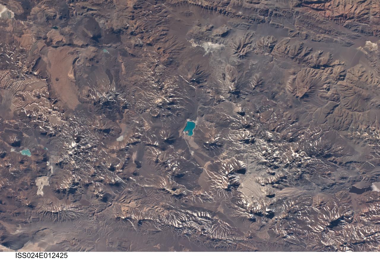 ISS024-E-012425 (22 Aug. 2010) --- This photograph, featuring a landscape in the central Andes mountains near the Chile/Argentina border dominated by numerous volcanoes and associated landforms, was photographed by an Expedition 24 crew member on the International Space Station. Layers of older sedimentary rocks are visible to the southeast (upper right). Many of the volcanic cones show grooves eroded by water to form gullies. Such erosion has occurred since the host volcano was built up, indicating that most volcanoes in this view have been inactive for centuries or millennia. A few volcanoes exhibit much less erosion, and even show tongues of recent, dark lava flows (top left). According to scientists, two of these volcanoes, Cerro el Condor and Peinado have likely erupted within approximately the last 12,000 years (the Holocene Epoch). Also visible in the image is the world’s highest active volcano, Nevado Ojos del Salado, with a summit at 6,887 meters above sea level. The most recent confirmed eruption of this volcano has been dated to 700 (approximately 300 years), but minor eruptive activity may have occurred as recently as 1993. Stratovolcanoes such as Cerro el Condor, Peinado, and Nevado Ojos del Salado are formed partly by buildup of lava flows and partly by buildup of explosively vented material dropping back down onto the surface. One type of material associated with explosive eruptions is welded tuff, which is formed by molten and fragmented rock that accumulates on the ground and later solidifies. A large tuff sheet is visible at top left. Formed very rapidly, these sheets have been termed “instant landscapes.” So active has the Andean volcanic system been that the origin of many of the tuffs in the Andes cannot be pinpointed since source vents have been overprinted by subsequent volcanic events. The volcanic landscape also shows that the erosive work of rivers—and glaciers repeatedly in the recent past—is slower than the opposite processes of the upward building of the volcanoes. The bright blue, nearly 7-kilometer-long lake near the center of the image is known as Laguna Verde. This and other less obvious lakes indicate that water (snowmelt or direct precipitation) is unable to reach the sea, but is rather impounded in the depressions between the volcanic edifices.