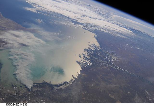 ISS024-E-014233 (11 Sept. 2010) --- A smoke plume near the northern Caspian Sea, Kazakhstan is featured in this image photographed by an Expedition 24 crew member on the International Space Station. This broad view of the north coast of the Caspian Sea shows a smoke plume (left) and two river deltas (bottom and lower right). The larger delta is that of the Volga River which appears prominently here in sunglint (light reflected off a water surface back towards the observer), and the smaller less prominent delta is that of the Ural River. Wide angle, oblique views ? taken looking outward at an angle, rather than straight down towards Earth ? such as this give an excellent impression of how crew members onboard the space station view Earth. For a sense of scale, the Caucasus Mts. (across the Caspian, top right) are approximately 1,100 kilometers to the southwest of the International Space Station?s nadir point location ? the point on Earth directly underneath the spacecraft ? at the time this image was taken. The smoke plume appears to be sourced in the dark-toned coastal marsh vegetation along the outer fringe of the Ural River delta, rather than in a city or at some oil storage facility. Although even small fires produce plumes that are long and bright and thus easily visible from space, the density of the smoke in this plume, and its 350-kilometer length across the entire north lobe of the Caspian Sea, suggest it was a significant fire. The smoke was thick enough nearer the source to cast shadows on the sea surface below. Lines mark three separate pulses of smoke, the most recent, nearest the source, extending directly south away from the coastline (lower left). With time, plumes become progressively more diffuse. The oldest pulse appears to be the thinnest, casting no obvious shadows (center left).