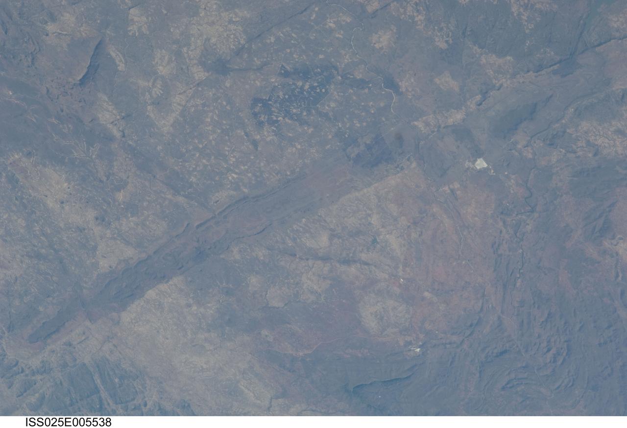 ISS025-E-005538 (30 Sept. 2010) --- The Great Dyke of Zimbabwe, Africa is featured in this image photographed by an Expedition 25 crew member on the International Space Station. The Great Dyke of Zimbabwe is a prominent geological feature that extends for over 550 kilometers, varying from 3-12 kilometers in width across the center of the country northeast – southwest; the southern end of the Dyke is illustrated in this view. The Dyke (or Dike in American English) is a layered mafic intrusion of igneous, metal-bearing rock that has been dated using uranium-lead isotopes to approximately 2.5 billion years in age, according to scientists. It intrudes even older rocks of the African craton, or core of oldest rocks forming the continent; in cross section, the Great Dyke looks somewhat triangular or keel-shaped suggesting to geologists that it rose along deep faults associated with extension of the African crust. Layered mafic intrusions are usually associated with economically important metals such as chromium, nickel, copper, platinum, titanium, iron, vanadium and tin. Chromium, in the form of the mineral chromite and platinum are particularly abundant in the Great Dyke and actively mined. Younger faults have offset sections of the Dyke along its length – two of the most obvious faults in the image are indicated, with arrows showing the relative directions of offset relative to the main trend of the intrusion. While the Great Dyke and its metal ores are products of geologic processes operating in the deep past, more recent events have also left their mark on the landscape as illustrated by two large fire burn scars which are visible at top center.
