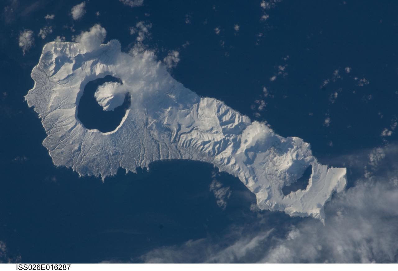 ISS026-E-016287 (9 Jan. 2011) --- Onekotan Island, part of the Russian Federation in the western Pacific Ocean, is featured in this image photographed by an Expedition 26 crew member on the International Space Station. Snow cover highlights calderas and volcanic cones that form the northern and southern ends of the island. Calderas are depressions formed when a volcano empties its magma chamber in an explosive eruption, followed by collapse of the overlaying material into the newly evacuated space. The northern end of the island is dominated by the Nemo Peak volcano that began forming within an older caldera approximately 9,500 years ago, according to scientists. The last recorded eruptive activity at Nemo Peak occurred in the early 18th century. The southern end of the island is formed by the 7.5 kilometer wide Tao-Rusyr Caldera. The caldera is filled by Kal’tsevoe Lake and Krenitzyn Peak, a volcano that has only erupted once in recorded history during 1952. Extending between northeastern Japan and the Kamchatka Peninsula of Russia, the Kuril Islands are an island arc located along the Pacific “Ring of Fire”. Island arcs form along an active boundary between two tectonic plates where one plate is being driven beneath the other (subduction). Magma generated by the subduction process feeds volcanoes—which eventually form volcanic islands—over the subduction boundary.