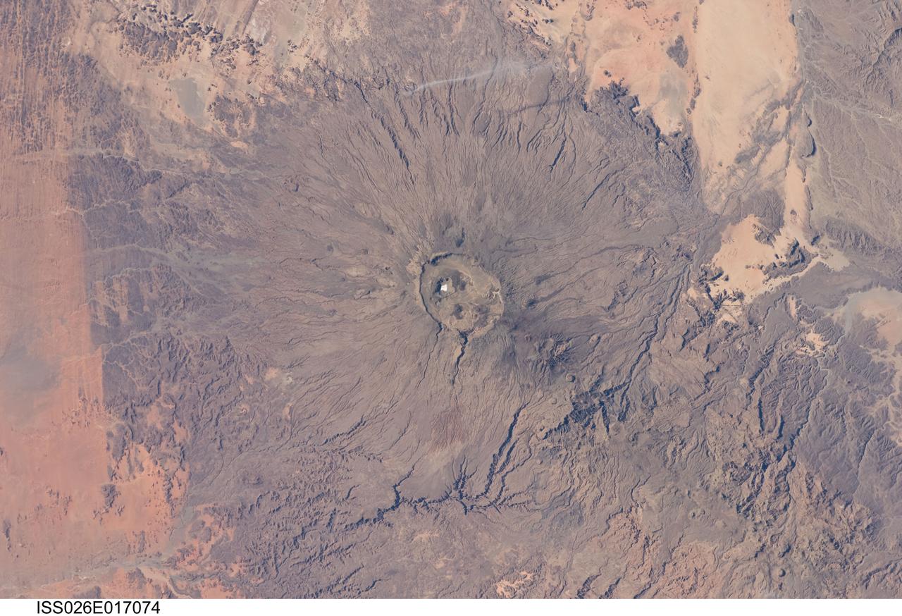 ISS026-E-017074 (11 Jan. 2011) --- Emi Koussi volcano in Chad is featured in this image photographed by an Expedition 26 crew member on the International Space Station. The large Emi Koussi volcano is located in northern Chad at the southeastern end of the Tibesti Range. The dark volcanic rocks of the volcano provide a sharp contrast to the underlying tan and light brown sandstones exposed to the west, south, and east. Emi Koussi is a shield volcano formed from relatively low viscosity lavas—flowing more like motor oil as opposed to toothpaste—and explosively-erupted ignimbrites that produce a characteristic low and broad structure that covers a wide area (approximately 60 x 80 kilometers). This photograph highlights the entire volcanic structure; at 3,415 meters above sea level, Emi Koussi is the highest summit of the Sahara region. The summit area contains three calderas formed by powerful eruptions. Two older, and overlapping, calderas form a depression approximately 12 x 15 kilometers in area bounded by a distinct rim (center). According to scientists, the youngest and smallest caldera, Era Kohor, formed as a result of eruptive activity that occurred within the past 2 million years. Young volcanic features including lava flows and scoria cones are also thought to be less than 2 million years old. There are no historical records of eruptive activity at Emi Koussi, but there is an active thermal area on the southern flank of the volcano.