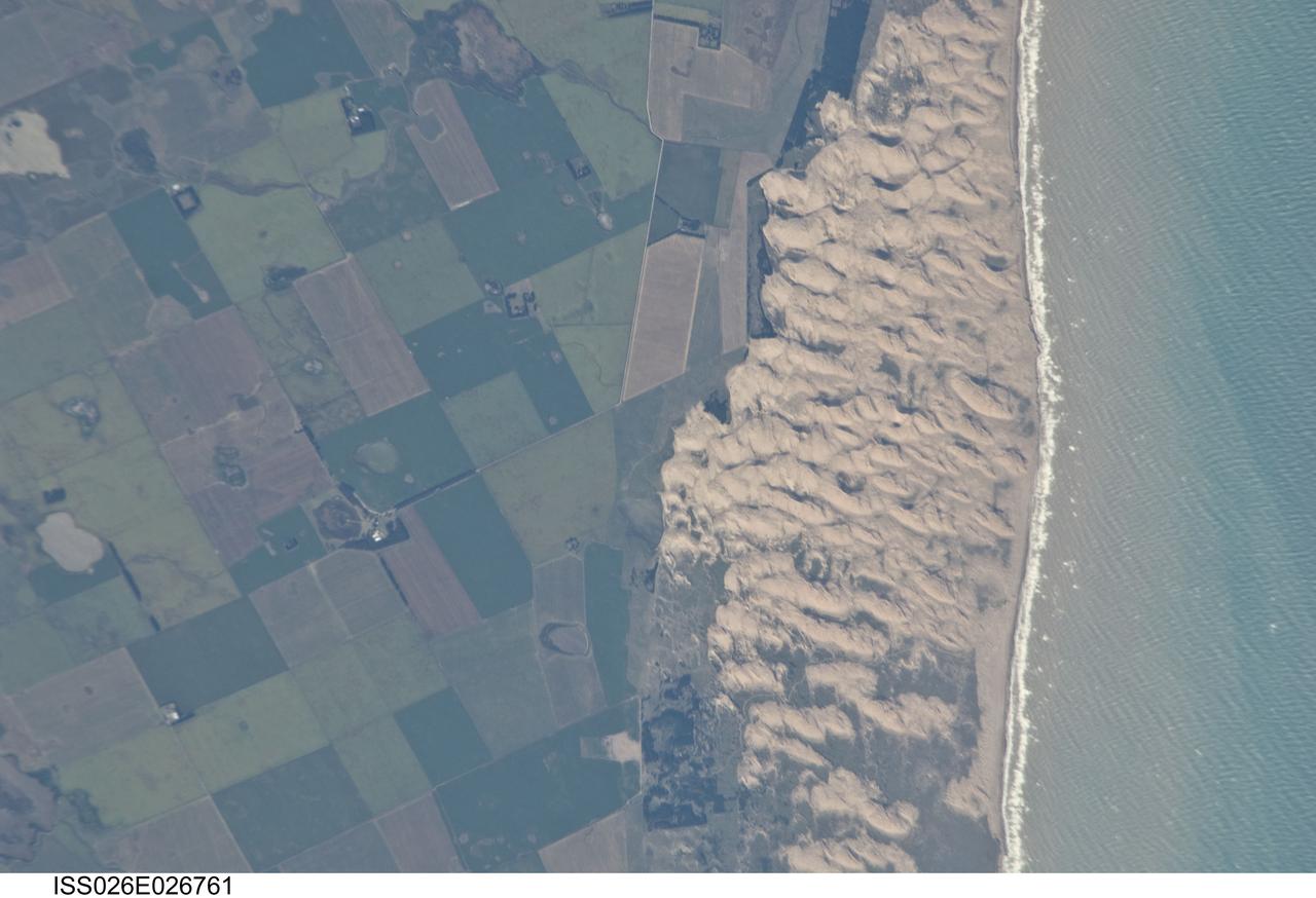 ISS026-E-026761 (13 Feb. 2011) --- Medano Blanco coastal dunes in Buenos Aires Province, Argentina are featured in this image photographed by an Expedition 26 crew member on the International Space Station. The Medano Blanco (White Dunes) are a well known recreational area 35 kilometers west of the twin cities of Nicochea-Quequen, one of the biggest ports in the province of Buenos Aires, central Argentina. The Medano Blanco dunes separate the Atlantic Ocean from intensive sunflower agricultural cropland, visible as the angular pattern of green and brown fields at left. The dune field extends 3.5 kilometers at its widest part. Narrow cordons of coastal dunes stretch along this part of Argentina’s coastline for hundreds of kilometers. Small streams, oriented towards the coast, cross the farmland at upper left. Water from these is dammed behind the dunes, and even within the dunes, where wetlands flourish. This is the drier southern part of Buenos Aires province near the more arid and windier border with Patagonia—one of the windiest places on Earth. The effect of strong westerly winds (blowing bottom to top in this slightly oblique image) can be seen everywhere in this detailed photograph. The dune sand is blown from local beaches onshore where it forms the dunes. The prominent dune ridge crests are oriented at right angles to the prevailing wind. Geologists suspect that erosion by wind has excavated numerous hollows which now dot the farmland as small shallow lakes. A stand of dark green trees has been planted on the upwind side of a health spa—located at the white dot (center left) which is the roof of a stately building—as a protective barrier against the wind. The white fringe along the seashore is formed from lines of breaking waves. Crossing swell trains can be seen offshore in the Atlantic Ocean. The curvature of the swell pattern offshore also shows the effect of the westerly wind, and indeed drives the local sea current in this area—slightly more prominent onshore swells approach the coast from the southeast. The light brown tint of seawater nearer the shore is the fine muddy fraction of the coastal sediment being washed seaward (after being stirred up by wave action) and then east by the current.