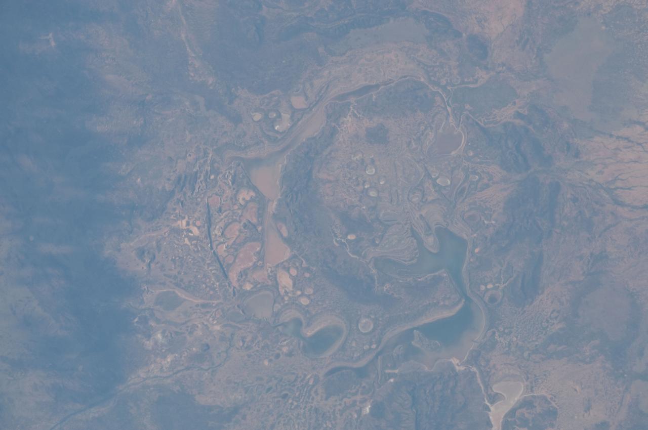 ISS028-E-014782 (6 July 2011) --- The Shoemaker (formerly Teague) Impact Structure, located in Western Australia in a drainage basin south of the Waldburg Range, presents an other-worldly appearance in this detailed photograph recorded from onboard the International Space Station on July 6. The Shoemaker impact site is approximately 30 kilometers in diameter, and is clearly defined by concentric ring structures formed in sedimentary rocks (brown to dark brown, image center) that were deformed by the impact event approximately 1630 million years ago, according to the Earth Impact Database. Several saline and ephemeral lakes?Nabberu, Teague, Shoemaker, and numerous smaller ponds?occupy the land surface between the concentric ring structures. Differences in color result from both water depth and suspended sediments, with some bright salt crusts visible around the edges of smaller ponds (image center The Teague Impact Structure was renamed Shoemaker in honor of the late Dr. Eugene M. Shoemaker, a pioneer in the field of impact crater studies and planetary geology, and founder of the Astrogeology Branch of the United States Geological Survey. The image was recorded with a digital still camera using a 200 mm lens, and is provided by the ISS Crew Earth Observations experiment and Image Science & Analysis Laboratory, Johnson Space Center.