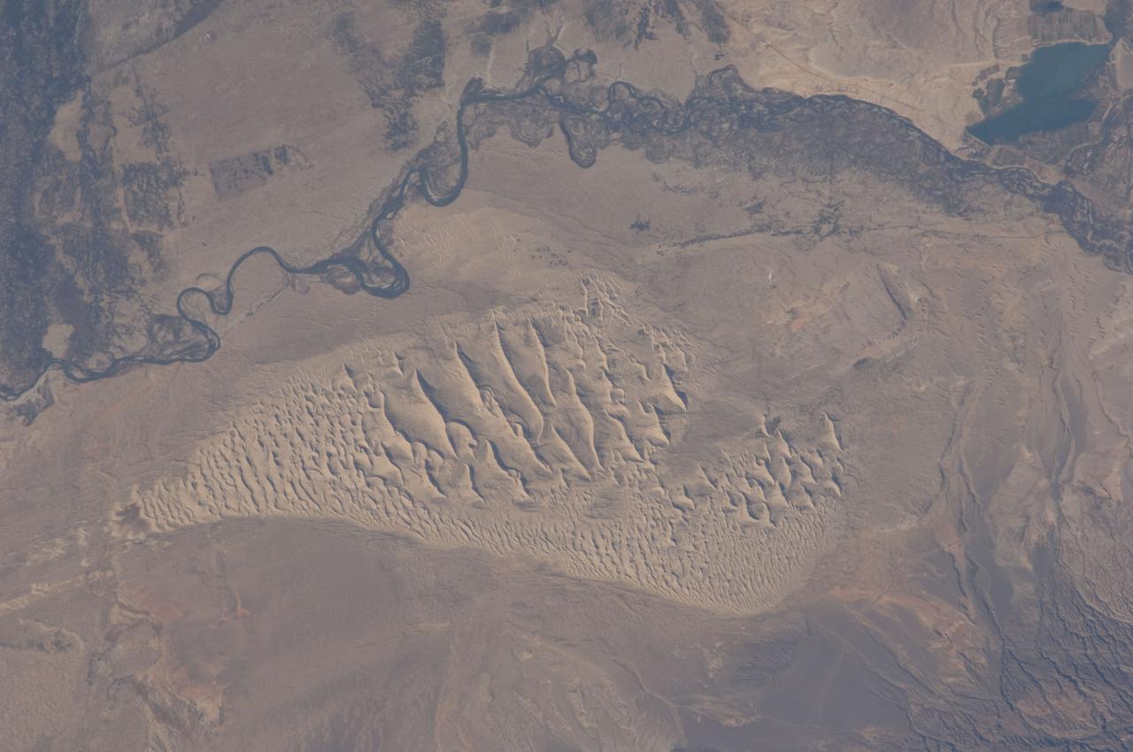 ISS028-E-044444 (8 Sept. 2011) --- This image, photographed by an Expedition 28 crew member on the International Space Station, highlights a sand dune field within the Burqin-Haba River-Jimunai Desert near the borders of China, Mongolia, Russia and Kazakhstan. The dune field (approximately 32 kilometers long) is located immediately to the west-northwest of the city of Burqin (not shown), and is part of the Junggar Basin, a region of active petroleum production in northwestern China. The Irtysh River, with associated wetlands and riparian vegetation (appearing grey-green in the image) flows from its headwaters in the Altay Mountains to the east towards Siberia to the west (right to left). Tan sandy linear dunes at center on the south side of the Irtysh River dominate the view. The linear dunes are formed from mobile barchan (crescent-shaped) dunes moving from left to right in this view; the barchans eventually merge to form the large linear dunes some of which reach 50-100 meters in height. Sand moving along the southern edge of the dune field appears to be feeding a southeastern lobe with a separate population of linear dunes (lower right). The Burqin-Haba River-Jimunai Desert area also includes darker gravel covered surfaces that form pavements known locally as gobi. These are somewhat indistinguishable from vegetated areas arresting some of the dunes at the resolution of the image, but tend to be located on the flat regions between the dunes.