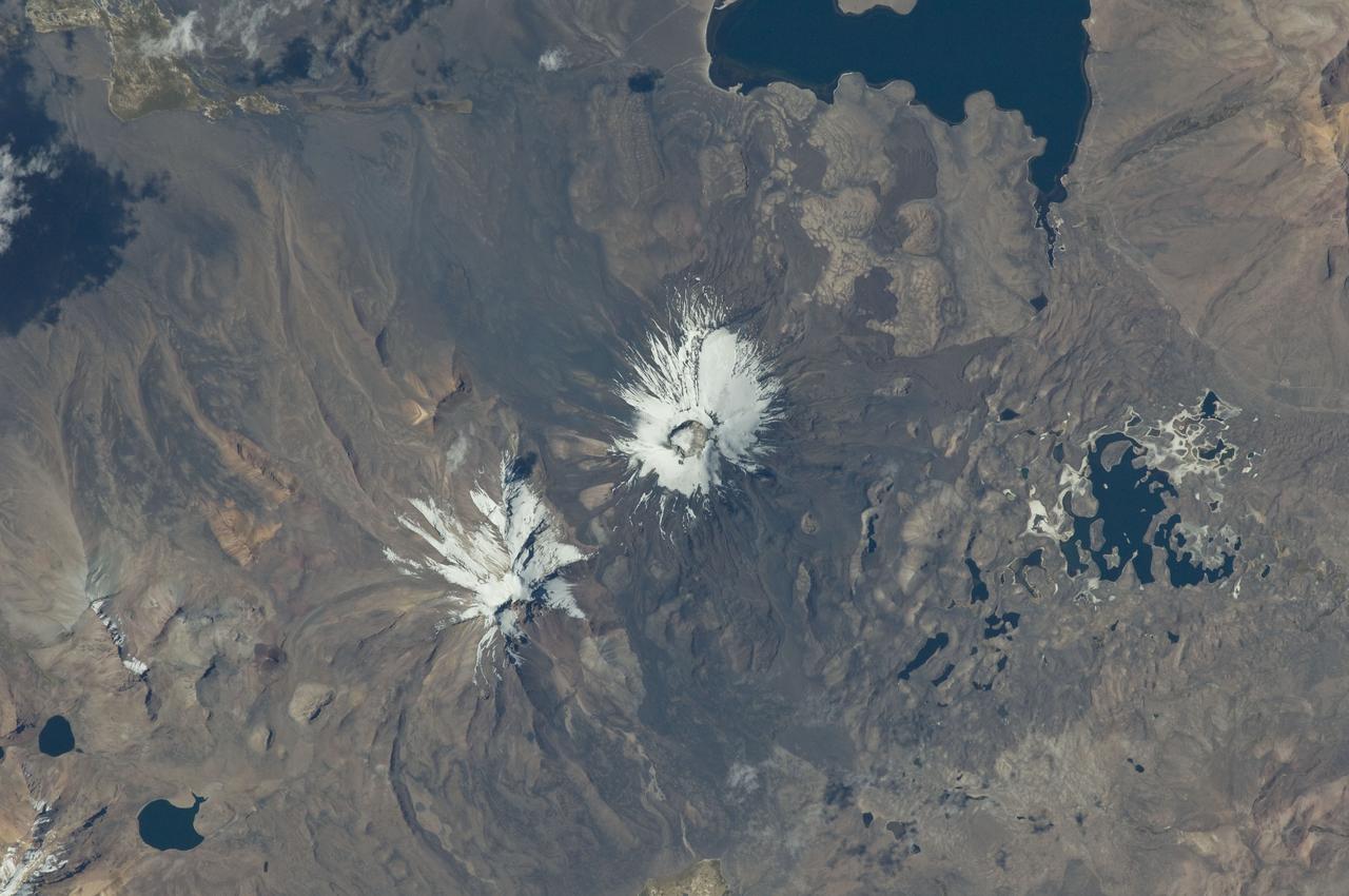 ISS029-E-020003 (7 Oct. 2011) --- Parinacota Volcano in the Chile-Bolivia border region, South America is featured in this image photographed by an Expedition 29 crew member on the International Space Station. Volcan Parinacota (?flamingo lake? in the regional Aymara language) is a potentially active stratovolcano located on the Altiplano, a high plateau situated within the Andes Mountains of west-central South America. While no direct observations of eruptive activity are recorded, surface exposure age-dating of lava flows suggests that activity occurred as recently as 290 AD approximately 300 years, according to scientists. Local Aymara stories also suggest that the volcano has erupted during the past 1,000 years. This detailed photograph highlights the symmetrical cone of Parinacota, with its well-developed summit crater (elevation 6,348 meters above sea level) at center. Dark brown to dark gray surfaces to the east and west of the summit include lava flows, pyroclastic deposits, and ash. A companion volcano, Pomerape, is located across a low saddle to the north ? scientists believe this volcano last erupted during the Pleistocene Epoch (extending from approximately 3 million to 12,000 years ago). The summits of both volcanoes are covered by white permanent snowpack and small glaciers. Together, the two volcanoes form the Nevados de Payachata volcanic area. Eruptive activity at Parinacota has directly influenced development of the local landscape beyond the emplacement of volcanic deposits ? approximately 8,000 years ago the western flank of the volcano collapsed, creating a debris avalanche that traveled 22 kilometers to the west. This debris avalanche blocked drainages, leading to the formation of Lake Chungara to the south (upper right). The uneven, hummocky surface of the debris avalanche deposit provides ample catchments for water, as evidenced by the numerous small ponds and Cotacotani Lake to the west.