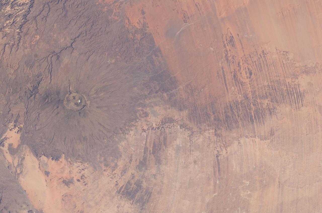 ISS030-E-005456 (26 Nov. 2011) --- Emi Koussi Volcano and Aorounga Impact Crater, Chad are featured in this image photographed by an Expedition 30 crew member on the International Space Station. This striking photograph features two examples of circular landscape features?labeled as craters?that were produced by very different geological processes. At left, the broad grey-green shield volcano of Emi Koussi is visible. The volcano is marked by three overlapping calderas formed by eruptions; these form a large oblong depression at the 3,415 meter ASL summit of the volcano.  A smaller crater sits within the larger caldera depression. While volcanic activity has not been observed, nor is mentioned in the historical record, an active thermal area is located on the southern flank. The circular Aorounga Impact Crater is located approximately 110 kilometers to the southeast of Emi Koussi and has its origin in forces from above rather than eruptions from below. According to scientists, the Aorounga structure is thought to record a meteor impact approximately 345-370 million years ago. The circular feature visible at upper right may be only one of three impact craters formed by the same event ? the other two are buried by sand deposits. The linear features (lower right) that arc around Emi Koussi and overprint Aorounga and the surrounding bedrock are known as yardangs; these are rock ridges formed by wind erosion.