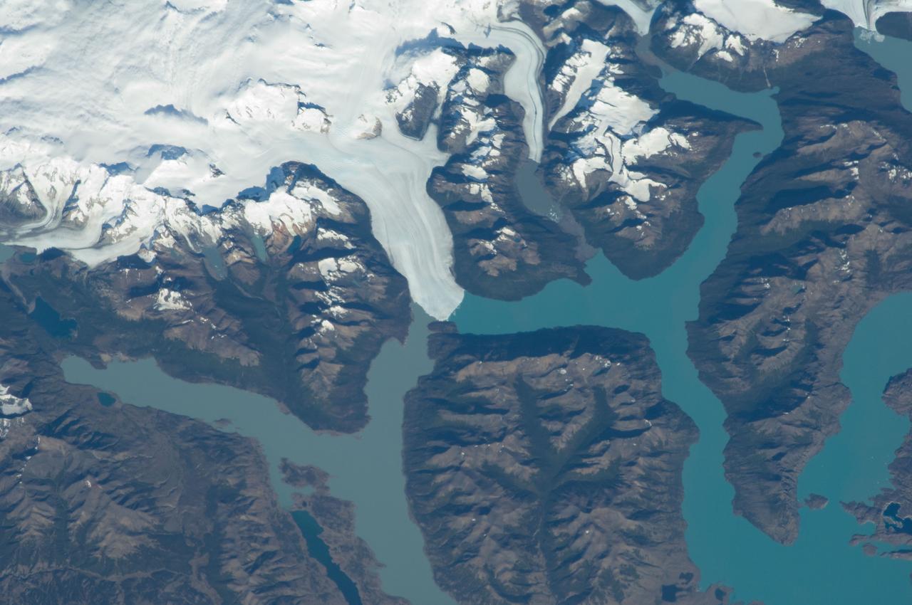 ISS030-E-091253 (21 Feb. 2012) --- Perito Moreno Glacier near Lake Argentino, Argentina is featured in this image photographed by an Expedition 30 crew member on the International Space Station. The largest glacier tongue in this image is known as the Perito Moreno Glacier (center).  It descends from the Southern Patagonian Icefield (top) at great altitudes (greater than 2,100 meters, 6,825 feet) in the southern Andes Mountains, down into the water and warmer altitudes of Lake Argentino at approximately 180 meters above sea level. The glacier is 30 kilometers long (image width represents approximately 60 kilometers on the ground). Perito Moreno is one of the largest glaciers in Patagonia, and is perhaps the most famous for the fact that it periodically cuts off the major southern arm (known as Brazo Rico) of Lake Argentino completely from the rest of the lake. This is because the glacier advances right across the lake until it meets the opposite shoreline. The ice tongue is “grounded” (meaning that it is not floating, as occurs at the termini of glaciers and ice shelves where they enter the sea), thus forming a natural dam which prevents the lake water on either side from circulating, which in turn causes muddier, “milkier” water to concentrate in Brazo Rico. Sub-ice water, flows under the ice, not only carrying the mud into the lake but also helping lubricate the glacier’s downhill movement. Because of its effect as a dam, meltwater from the south raises water levels in Brazo Rico by as much as 30 meters above the level of the water in Lago Argentino. The great pressure of this higher water ultimately causes the ice tongue to rupture catastrophically, in a great natural spectacle. The last rupture took place in March 2012. The process then repeats, on average every four to five years, as the glacier starts to grow back towards the opposite shoreline. The repeatability of the rupture has contributed to the event becoming a major tourist attraction in the region.