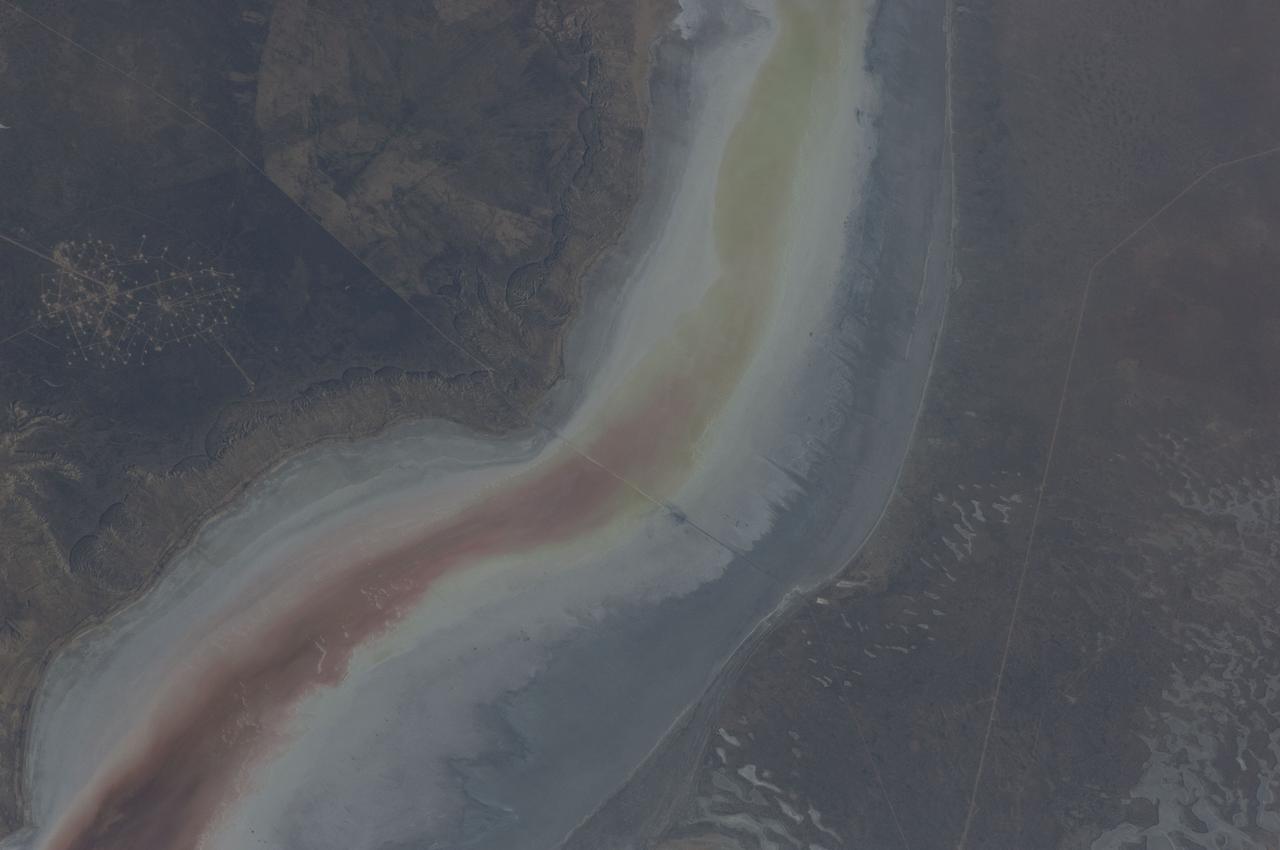 ISS031-E-030896 (11 May 2012) --- The Sor Kaydak near the northeast Caspian Sea in Kazakhstan are featured in this image photographed by an Expedition 31 crew member on the International Space Station. This striking image shows saline water of different colors in the Sor Kaydak, a salt marsh that leads into the northeast bulb of the Caspian Sea. The central 50 kilometers of this 180 kilometer-long depression is shown in this photograph. The Sor Kaydak depression is inundated on occasion by water from the Caspian Sea since it lies at the same elevation?that is, 29 meters below global sea level?though separated from the Sea by a low bar of land that is 1?2 meters high. The different colors of marsh water (brown to pink to light green, northeast to southwest) result from the interplay of water depth and resident organisms such as algae. Algae color varies depending on water temperature and salinity. The irregular gray areas at bottom right are wet zones between low sand dunes. These interdune flats are whitened with salt derived by evaporation of Caspian Sea water (the sea is just outside the image bottom right). The jagged line following the colored water (crossing the center of the image top to bottom) is the limit of the wetting zone (or perimeter), an irregular zone influenced by wind and the depth of water in the marsh. Small cliffs can be seen marking the east margin of the depression that contains Sor Kaydak. Above the cliffs a plateau surface (approximately 200 meters above the salt marsh, approximately 160 meters above global sea level) extends eastward for hundreds of kilometers. Here the plateau is occupied by a dense pattern of well heads which appear as a geometric pattern of tan dots. By contrast, the west margin (right) rises less than 10 meters above the marsh altitude. The straight line visible at center is a pipeline built across the salt marsh which takes oil to a terminal on the Caspian shore 100 kilometers northwest of the area shown here.