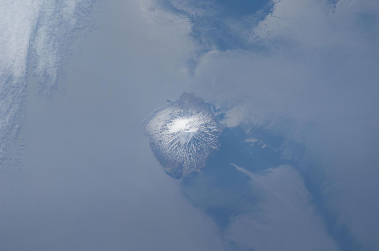 ISS031-E-041959 (18 May 2012) --- Alaid Volcano in the Kuril Islands of the Russian Federation is featured in this image photographed by an Expedition 31 crew member on the International Space Station. The Kurils chain extends from the Kamchatka Peninsula to the islands of Japan, and contains numerous active volcanoes along its length. Alaid is the highest (2,339 meters above sea level) volcano in the Kuril chain, as well as being the northernmost. The textbook conic morphology of this stratovolcano is marred only by the summit crater, which is breached to the south (center) and highlighted by snow cover. The volcano rises 3,000 meters directly from the floor of the Sea of Okhotsk, with the uppermost part of the volcanic edifice exposed as an island. Much of the sea surface surrounding the volcano has a silver-gray appearance. This mirror-like appearance is due to sunglint, where light reflects off the sea surface and is scattered directly towards the observer onboard the space station. Sunglint is largely absent from a zone directly to the west of the volcano, most likely due to surface wind or water current patterns that change the roughness?and light scattering properties?of the water surface in this area. Volcanoes in the Kurils, and similar island arcs in the Pacific ?ring of fire?, are fed by magma generated along the boundary between two tectonic plates, where one plate is being driven beneath the other (a process known as subduction). Alaid Volcano has been historically active with the most recent confirmed explosive activity occurring in 1996.