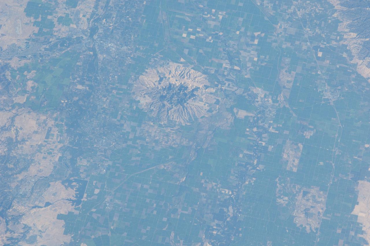 ISS032-E-010482 (29 July 2012) --- Sutter Buttes in California are featured in this image photographed by an Expedition 32 crew member on the International Space Station. Sometimes called the ?smallest mountain range in the world?, the Sutter Buttes rise almost 610 meters above the surrounding flat agricultural fields of the Great Valley of central California. Scientists believe the Sutter Buttes are remnants of a volcano that was active approximately 1.6 ? 1.4 million years ago during the Pleistocene Epoch. The central core of the Buttes is characterized by lava domes?piles of viscous lava that erupted onto the surface, building higher with each successive layer. Today, these lava domes form the high central hills of the Buttes; shadows cast by the hills are visible at center. Surrounding the central core is an apron of fragmental material created by occasional eruptions of the lava domes ? this apron extends roughly 18 kilometers east-west and 16 kilometers north-south. The volcanic material was transported outwards from the central core during eruptions by hot gasses (pyroclastic flows) or by cooler water-driven flows (lahars). Later stream erosion of the debris apron is evident from the radial drainage pattern surrounding the central core. A third geomorphic region of valleys known as the ?moat? is present between the core and the debris apron, and was formed from erosion of older, exposed sedimentary rocks that underlie the volcanic rocks. The Sutter Buttes present a striking visual contrast with the surrounding green agricultural fields?here mostly rice, with some sunflower, winter wheat, tomato, and almonds?of the Great Valley. Urban areas such as Yuba City, CA (located 18 kilometers to the southeast) appear as light to dark gray stippled regions. Sacramento, CA (not shown) is located approximately 80 kilometers to the south-southeast. The image appears slightly distorted (oblique) due to the viewing angle from the space station.