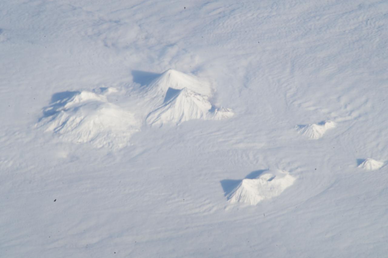 ISS033-E-018010 (3 Nov. 2012) --- Volcanoes in central Kamchatka are featured in this image photographed by an Expedition 33 crew member on the International Space Station. The snow-covered peaks of several volcanoes of the central Kamchatka Peninsula are visible standing above a fairly uniform cloud deck that obscures the surrounding lowlands. In addition to the rippled cloud patterns caused by interactions of air currents and the volcanoes, a steam and ash plume is visible at center extending north-northeast from the relatively low summit (2,882 meters above sea level) of Bezymianny volcano. Volcanic activity in this part of Russia is relatively frequent, and well monitored by Russia’s Kamchatka Volcanic Eruption Response Team (KVERT). The KVERT website provides updated information about the activity levels on the peninsula, including aviation alerts and webcams. Directly to the north and northeast of Bezymianny, the much larger and taller stratovolcanoes Kamen (4,585 meters above sea level) and Kliuchevskoi (4,835 meters above sea level) are visible. Kliuchevskoi, Kamchatka’s most active volcano, last erupted in 2011 whereas neighboring Kamen has not erupted during the recorded history of the region. An explosive eruption from the summit of the large volcanic massif of Ushkovsky (3,943 meters above sea level; left) northwest of Bezymianny occurred in 1890; this is the most recent activity at this volcano. To the south of Bezymianny, the peaks of Zimina (3,081 meters above sea level) and Udina (2,923 meters above sea level) volcanoes are just visible above the cloud deck; no historical eruptions are known from either volcanic center. While the large Tobalchik volcano to the southwest (bottom center) is largely formed from a basaltic shield volcano, its highest peak (3,682 meters above sea level) is formed from an older stratovolcano. Tobalchik last erupted in 1976. While this image may look like it was taken from the normal altitude of a passenger jet, the space station was located approximately 417 kilometers above the southeastern Sea of Okhotsk; projected downwards to Earth’s surface, the space station was located over 700 kilometers to the southwest of the volcanoes in the image. The combination of low viewing angle from the orbital outpost, shadows, and height and distance from the volcanoes contributes to the appearance of topographic relief visible in the image.