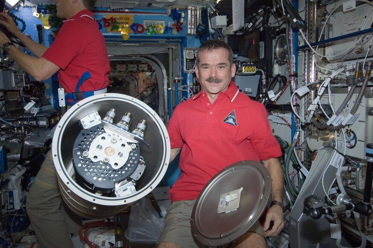 View of Canada Space Agency (CSA) Chris Hadfield, Expedition 34 Flight Engineer (FE), poses with a Materials Science Laboratory (MSL) Furnace Launch Support Structure (FLSS) in the U.S. Laboratory.  Tom Marshburn (background), Expedition 34 FE uses laptop computer.  Photo was taken during Expedition 34.