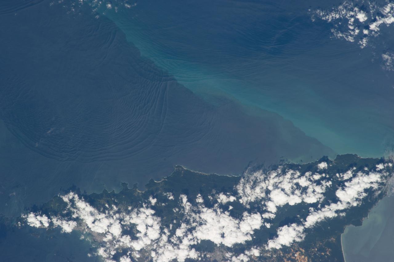 ISS034-E-032377 (18 Jan. 2013) --- Internal waves off Northern Trinidad are featured in this image photographed by an Expedition 34 crew member on the International Space Station. This photograph shows the north coast of the island of Trinidad in the southeastern Caribbean Sea, where heating of the land is setting off the growth of cumulus clouds. The light blue northwest-southeast trending plume at center is sediment from one of the rivers that flows into the sea here. Adjacent to, and appearing to cross the sediment plume, a series of subtle interacting arcs can be seen in the sea. These are known as internal waves which are the surface manifestation of slow waves moving tens of meters beneath the sea surface. These produce enough of an effect on the sea surface to be seen from space, but only where they are enhanced due to reflection of sunlight, or sunglint, back towards the space station. The image shows at least three sets of internal waves interacting. The most prominent set (top left) shows a “packet” of several waves moving from the northwest due to the tidal flow towards the north coast of Trinidad. Two less prominent, younger sets can be seen further out to sea. A very broad set enters the view from the north and northeast, and interacts at top center with the first set. All the internal waves are probably caused by the shelf break near Tobago (outside the image to top right). The shelf break is the step between shallow seas (around continents and islands) and the deep ocean. It is the line at which tides usually start to generate internal waves. The sediment plume at center is embedded in the Equatorial Current (also known as the Guyana Current) and is transporting material to the northwest—in almost the opposite direction to the movement of the internal waves. The current flows strongly from east to west around Trinidad, all the way from equatorial Africa, driven by year-round easterly winds. Seafarers in the vicinity of Trinidad are warned that the current, and its local reverse eddies, make navigation of smaller craft in these waters complicated and sometimes dangerous.