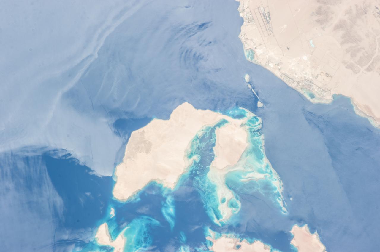 ISS036-E-010628 (24 June 2013) --- Strait of Tiran, Red Sea and Gulf of Aqaba are featured in this image photographed by an Expedition 36 crew member on the International Space Station. The approximately six-kilometer wide Strait of Tiran (also known as the Straits of Tiran) between the Egyptian mainland and Tiran Island separates the Gulf of Aqaba from the Red Sea, and provides two channels (290 meters and 73 meters deep, respectively) navigable by large ships bound for ports in Jordan and Israel. A smaller passage also exists between the east side of Tiran Island and Saudi Arabia, but this a single channel that is 16 meters deep. Due to its strategic position, control of the Strait has been an important factor in historical conflicts of the region, such as the Suez Crisis in 1956 and the Six-Day War in 1967. This photograph illustrates the morphology of the Strait. The relatively clear, deep-water passages of the western Strait of Tiran are visible at right, while the more sinuous shallow-water passage on the Saudi Arabia side can be seen at bottom center. Light blue to turquoise areas around Tiran Island indicate shallow water, while the island itself is arid and largely free of vegetation. Coral reefs are also found in the Straits of Tiran and are a popular diving destination. The silvery sheen on the water surface within the Strait and the south of Tiran Island is sunglint – light reflecting off the water surface back towards the observer on the space station. Disturbance to the water surface, as well as presence of substances such as oils and surfactants, can change the reflective properties of the water surface and highlight both surface waves and subsurface currents. For example, a large wave set is highlighted by sunglint at upper left.