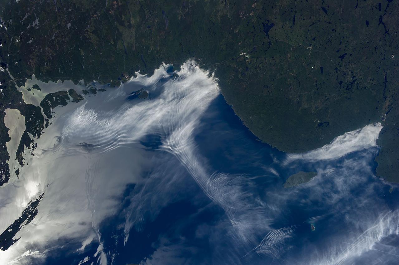 ISS036-E-011843 (24 June 2013) --- Gravity waves and sunglint on Lake Superior are featured in this image photographed by an Expedition 36 crew member on the International Space Station. From the vantage point of the space station, crew members frequently observe Earth atmospheric and surface phenomena in ways impossible to view from the ground. Two such phenomena?gravity waves and sunglint?are illustrated in this photograph of northeastern Lake Superior. The Canadian Shield of southern Ontario (bottom) is covered with extensive green forest canopy typical of early summer. Offshore, and to the west and southwest of Pukaskwa National Park several distinct sets of parallel cloud bands are visible. Gravity waves are produced when moisture-laden air encounters imbalances in air density, such as might be expected when cool air flows over warmer air; this can cause the flowing air to oscillate up and down as it moves, causing clouds to condense as the air rises (cools) and evaporate away as the air sinks (warms). This produces parallel bands of clouds oriented perpendicular to the wind direction. The orientation of the cloud bands visible in this image, parallel to the coastlines, suggests that air flowing off of the land surfaces to the north is interacting with moist, stable air over the lake surface, creating gravity waves. The second phenomenon?sunglint?effects the water surface around and to the northeast of Isle Royale (upper right). Sunglint is caused by light reflection off a water surface; some of the reflected light travels directly back towards the observer, resulting in a bright mirror-like appearance over large expanses of water. Water currents and changes in surface tension (typically caused by presence of oils or surfactants) alter the reflective properties of the water, and can be highlighted by sunglint. For example, surface water currents are visible to the east of Isle Royale that are oriented similarly to the gravity waves ? suggesting that they too are the product of winds moving off of the land surface.