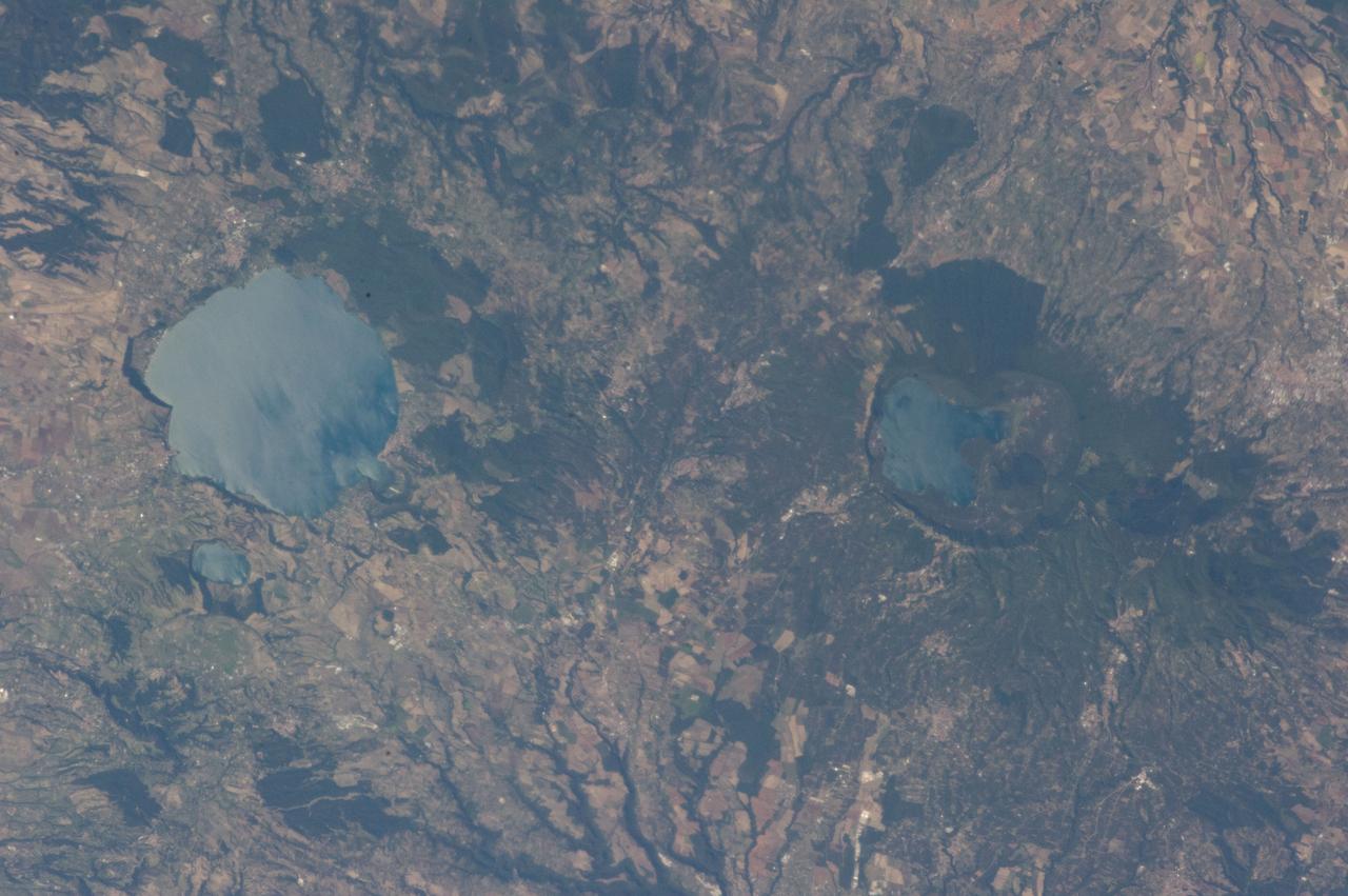 ISS036-E-039778 (3 Sept. 2013) --- Caldera lakes to the northwest of Rome, Italy are featured in this image photographed by an Expedition 36 crew member on the International Space Station. The Lazio region of central Italy has many landforms of volcanic origin, including several large lakes that mark the locations of ancient volcanoes. This photograph highlights two such lakes, Lago di Vico and Lago Bracciano, located to the northwest of the capital city of Rome. Both lakes are located within calderas, large depressions that form after violent explosive eruptions empty a volcano’s underlying magma chamber. Any remnants of the volcanic edifice can then collapse into the newly-formed void space, leading to the creation of large depressions. These depressions can then fill partially or completely with water, forming permanent lakes. Lago Bracciano (left) is the larger of the two lakes highlighted in the image; it is approximately eight kilometers wide at its widest point, and is located 32 kilometers northwest of Rome. According to scientists, the volcanic activity that led to the formation of Lago Bracciano began approximately 600,000 years ago and continued to approximately 40,000 years ago as part of the formation of the Sabatini volcanic complex. While part of the lake formation was due to caldera collapse of part of a large magma chamber, the current depression was also formed by movement along numerous faults in the area – a process known as volcano-tectonic collapse. Located approximately 24 kilometers to the north-northwest of Lago Bracciano, Lago di Vico (right) occupies part of a caldera associated with eruptive activity that began approximately 800,000 years ago and continued until approximately 90,000 years ago. The caldera formed largely by the catastrophic eruption of the ancestral Vico volcano approximately 200,000-150,000 years ago. The final phase of volcanic activity in the caldera led to the formation of a small lava cone in the northeast quadrant known as Mount Venus. The extent of the lakes of Bracciano and Vico are readily apparent in this image due to sunglint – light reflecting back towards the observer from the water surfaces. This reflection gives a mirror-like sheen to the water surfaces in the image. Dark green forested areas associated with parks are visible near both lakes, while light gray to white regions indicate built areas - such as the city of Viterbo at right - and tilled fields (bottom center).