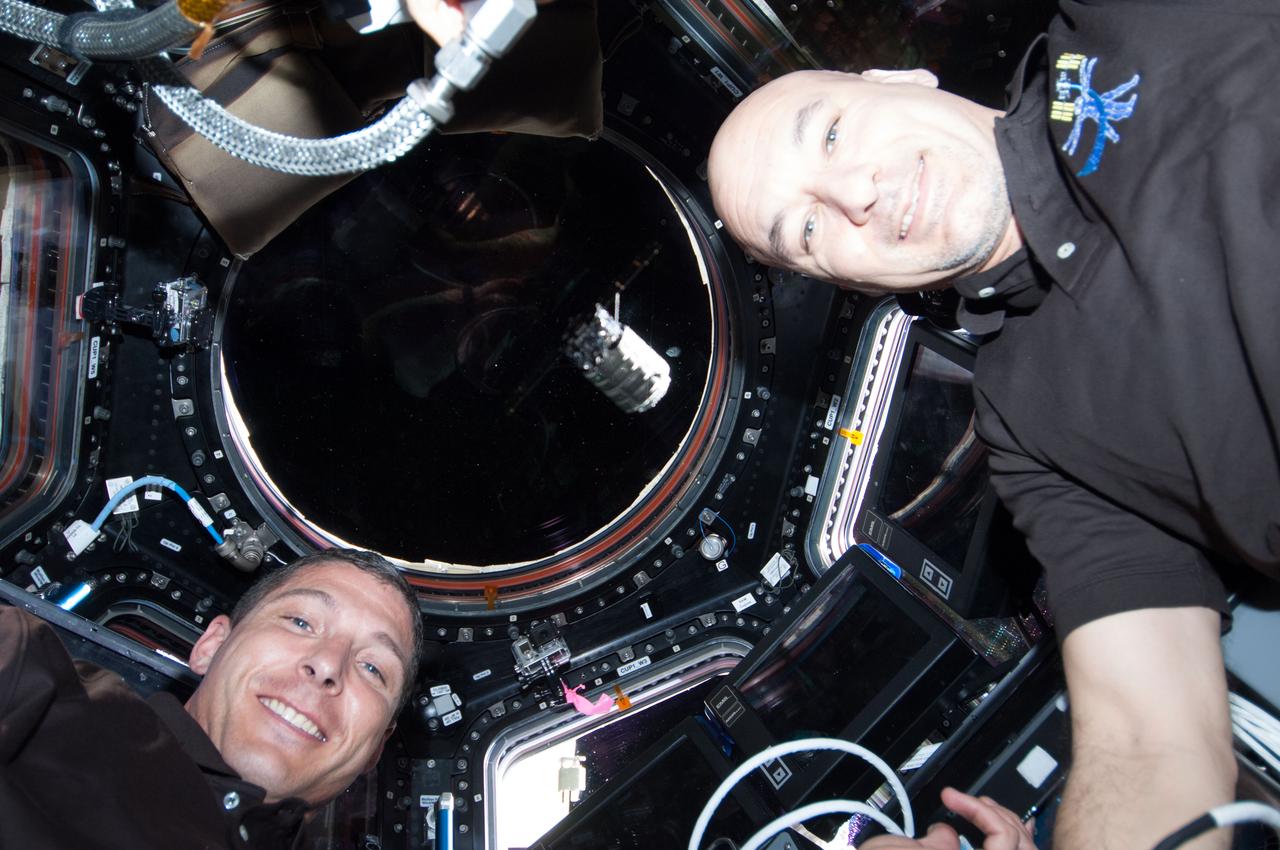 ISS037-E-003935 (29 Sept. 2013) --- NASA astronaut Michael Hopkins (left) and European Space Agency astronaut Luca Parmitano, both Expedition 37 flight engineers, pose for a photo near the windows in the International Space Station?s Cupola during rendezvous, capture and docking operations with the first Cygnus commercial cargo spacecraft (visible at center frame) built by Orbital Sciences Corp. The two spacecraft converged at 7:01 a.m. EDT on Sept. 29, 2013.