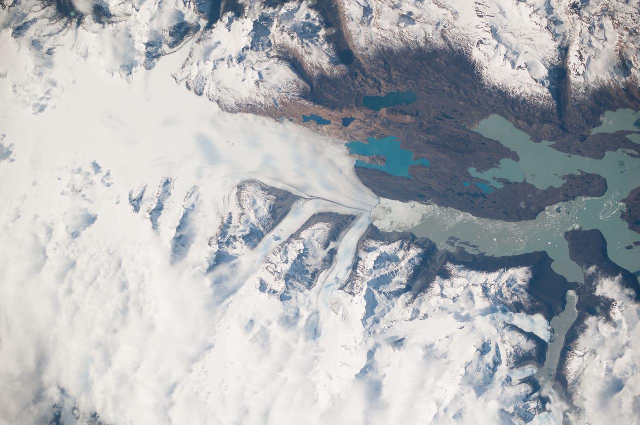 ISS037-E-005104 (2 Oct. 2013) --- Upsala Glacier Retreat and Patagonia Icefield are featured in this image photographed by an Expedition 37 crew member on the International Space Station. This photograph highlights the snout of the Upsala Glacier (49.88S 73.3W) on the Argentine side of the North Patagonian Icefield.  Ice flow in this glacier (white mass, left) is from the north (left).  Dark lines of rocky moraine within the ice give a sense of the slow ice flow from left to right. A smaller side glacier joins Upsala at the present-day ice front—the wall from which masses of ice periodically collapse into Lake Argentino. In this image the 2.75-kilometer-long wall casts a thin, dark shadow. The surface of Lake Argentino is whitened by a mass of ice debris from a recent collapse of the ice wall. Larger icebergs that have calved appear as white dots on the lake surface at right. Remotely sensed data, including detailed astronaut images such as this, have recorded the position of the ice front over the years.  Even though the ice actually flows slowly southward, comparison of this October 2013 image with older data (not shown) indicates that the ice wall of the glacier has moved backwards—upstream—an average of 3.6 kilometers since early 2002. This so-called “retreat” is believed by scientists to indicate local climatic warming in this part of South America. The warming not only causes the ice front to retreat but more importantly, causes overall thinning of the glacier ice mass, as a study of 63 glaciers in Patagonia has shown is now a general trend (Rignot et al. 2003). Ice-front retreat is now known to be related to volumetric loss due to melting. Water color is related to glacier flow.  Lake Argentino receives most of the ice from the glacier and thus also receives most of the “rock flour” (rocks ground to white powder by the ice scraping against the rock floor of the valley) from underneath the glacier. Glacial flour turns the lake water a gray-green hue in this image. The darker blue of the smaller lakes (top) indicates that they are receiving much less rock flour.