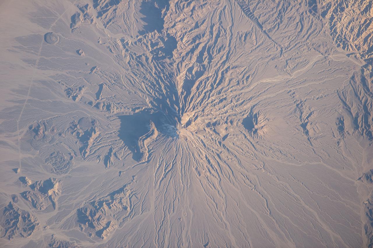 ISS038-E-025895 (5 Jan. 2014) --- Bazman volcano in Iran is featured in this image photographed by an Expedition 38 crew member on the International Space Station. Bazman volcano is located in a remote southern region within the Bazman Protected Area of Sistan and Baluchestan Provinces. While the volcano has the classic cone shape associated with stratovolcanoes, it is also heavily dissected by channels that extend downwards from the 3,490-meter-above-sea-level summit. This radial drainage pattern - looking similar to the spokes of a bicycle wheel - is readily observed in this photograph. Such patterns can form around high, symmetric peaks when water runoff and erosion is not constrained by the resistance of geologic materials or barriers to flow, leading to essentially even distribution of water runoff channels around the central peak. While there is no historical record of volcanism at Bazman, and no geologic record of eruptive activity within the past 10,000 years, some fumarolic activity - gas and steam emissions - have been reported, according to the Smithsonian Institution National Museum of Natural History's Global Volcanism Program. The summit of the volcano is marked by a well-formed explosion crater, and lava cones formed on the flanks of the main volcano are associated with well-preserved lava flows-a particularly striking example is visible on the north flank of Bazman at center. Together, these observations and features are suggestive that Bazman may be a dormant, rather than extinct, volcano.