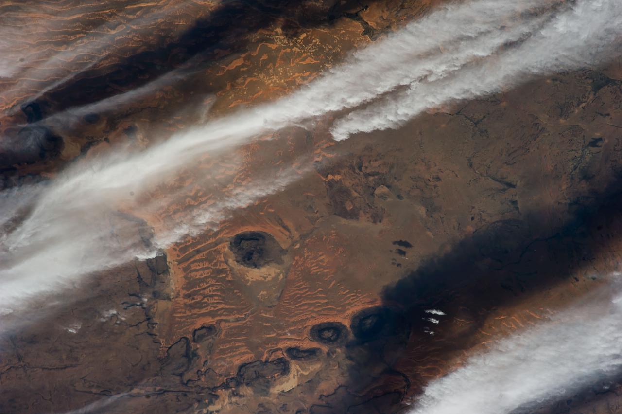ISS038-E-026862 (8 Jan. 2014) --- The Western Sahara Desert is featured in this image photographed by an Expedition 38 crew member on the International Space Station. The infrequent cloud bands over southern Mauritania were photographed with an oblique look angle so that the dark cloud shadows are also a prominent part of the view. Beneath the cloud the Aouker plateau of dark sedimentary rocks appears as a ragged, near-vertical escarpment at top right. Isolated remnants of the plateau appear as dark mesas (flat-topped hills) across the top and near the center of the image. The escarpment is about 250 meters high, with a field of orange-colored dunes at the base (upper right). Prevailing winds in this part of the Sahara Desert blow from the northeast. The wavy dunes are aligned transverse to these winds. The sand that makes the dunes is blown in from a zone immediately upwind (just out of the bottom of the image) where dry river beds and dry lakes provide large quantities of mobile sand for the wind to transport. This pattern is typical in the western Sahara Desert, where plateau surfaces are mostly dune free and dune fields are located in the surrounding lowlands where the larger rivers deposit quantities of sandy sediment on the few occasions when they flow-sometimes only once in decades.