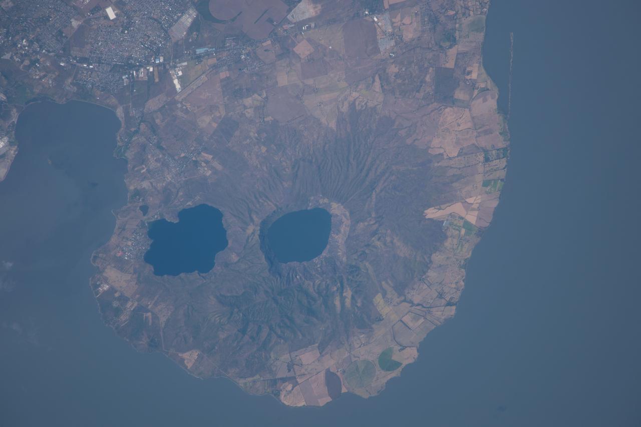 ISS038-E-035123 (21 Jan. 2014) --- Apoyeque Volcano, Nicaragua is featured in this image photographed by an Expedition 38 crew member on the International Space Station. The Chiltepe Peninsula, highlighted in this photograph, extends into Lake Managua in west-central Nicaragua. The peninsula is formed from part of a large ignimbrite shield, a geologic structure created by deposition of primarily low density materials (such as pumice) ejected during violent, explosive eruptive activity. Ignimbrite deposits are most commonly emplaced during large pyroclastic flows - gravity-driven mixtures of rock, ash, and volcanic gases that can cover hundreds of kilometers at speeds of hundreds of kilometers per hour - with ignimbrite shields formed over geologic time by successive flows. The Apoyeque caldera, filled with a 2.8-kilometer-wide and 400-meter-deep lake, dominates the center of the peninsula. Geological evidence indicates that Apoyeque last erupted around 50 BCE (plus or minus 100 years). The Laguna Xiloa maar - a volcanic crater formed by the explosive interaction of magma and groundwater - is located immediately to the southeast of Apoyeque and is also filled with a lake. According to scientists, Laguna Xiloa last erupted approximately 6,100 years ago. More recently, a swarm of small earthquakes was detected near Apoyeque in 2012. These seismic swarms, when detected in volcanically active areas, may indicate movement of magma prior to an eruption. The capital city of Managua, not visible in the image, is located approximately 15 kilometers to the southeast of Apoyeque, while the town of Bosques de Xiloa is considerably closer (approximately four kilometers).
