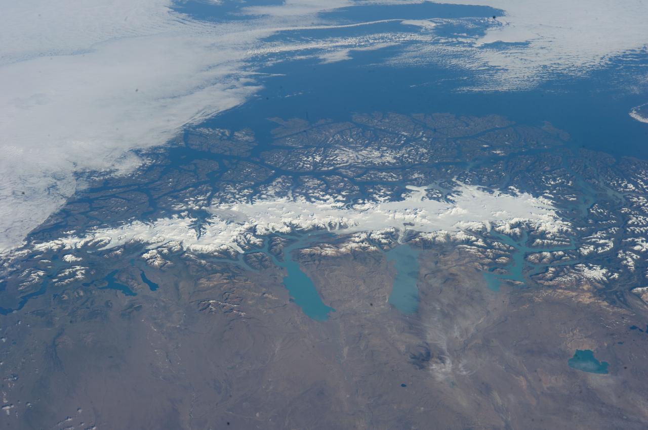 ISS038-E-047324 (13 Feb. 2014) --- This grand panorama of the Southern Patagonia Icefield (center) was imaged by an Expedition 38 crew member on the International Space Station on one of the rare clear days in the southern Andes Mountains. With an area of 13,000 square kilometers, the icefield is the largest temperate ice sheet in the Southern Hemisphere. Storms that swirl into the region from the southern Pacific Ocean (top) bring rain and snow (equivalent to a total of 2-11 meters of rainfall per year) resulting in the buildup of the ice sheet shown here (center). During the ice ages the glaciers were far larger. Geologists now know that ice tongues extended far onto the plains in the foreground, completely filling the great Patagonian lakes on repeated occasions. Similarly, ice tongues extended into the dense network of fjords (arms of the sea) on the Pacific side of the icefield. Ice tongues today appear tiny compared to the view that an "ice age" astronaut would have seen. A study of the surface topography of sixty-three glaciers, based on Shuttle Radar Topography Mission (SRTM) data, compared data from 2000 to data from studies going back about 30 years (1968-1975). Many glacier tongues showed significant annual "retreat" of their ice fronts, a familiar signal of climate change. The study also revealed that the almost invisible loss by glacier thinning is far more significant in explaining ice loss. The researchers concluded that volume loss by frontal collapse is 4-10 times smaller than that caused by thinning. Scaled over the entire icefield, including frontal loss (so-called calving when ice masses collapse into the lakes), it was calculated that 13.5 cubic kilometers of ice was lost each year over the study period. This number becomes more meaningful compared with the rate measured in the last five years of the study (1995-2000), when the rate increased almost threefold, averaging 38.7 cubic kilometers per year.  Extrapolating results from the low altitude glacier tongues implies that the high plateau ice on the spine of the Andes is thinning as well. In the decade since this study the often-imaged Upsala Glacier has retreated a further three kilometers, as shown recently in images taken by crew members aboard the space station. Glacier Pio X, named for Pope Pius X, is the only large glacier that is growing in length.