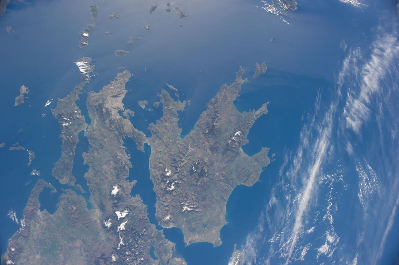 ISS039-E-003505 (21 March 2014) --- The Peloponnese, Greece is featured in this image photographed by an Expedition 39 crew member on the International Space Station. This view shows most of Greece. The urban region of Athens is readily recognizable due to its size and light tone compared to the surrounding landscape, although smaller Megara and Lamia also stand out. The dark-toned mountains with snow-covered peaks contrast with the warmer, greener valleys where agriculture takes place. The intense blue of the Mediterranean Sea fades near the sun?s reflection point, where numerous wind streaks in the lee of the island become visible along the right side of the image. The Peloponnese, ancient Sparta, is the great peninsula separated from the mainland by the narrow Isthmus of Corinth. Several times over the centuries the narrows have acted as a defensive point against attack from the mainland. More recently in 1893 the narrows provided a point of connection when a ship canal was excavated between the gulfs to the west and to the east.