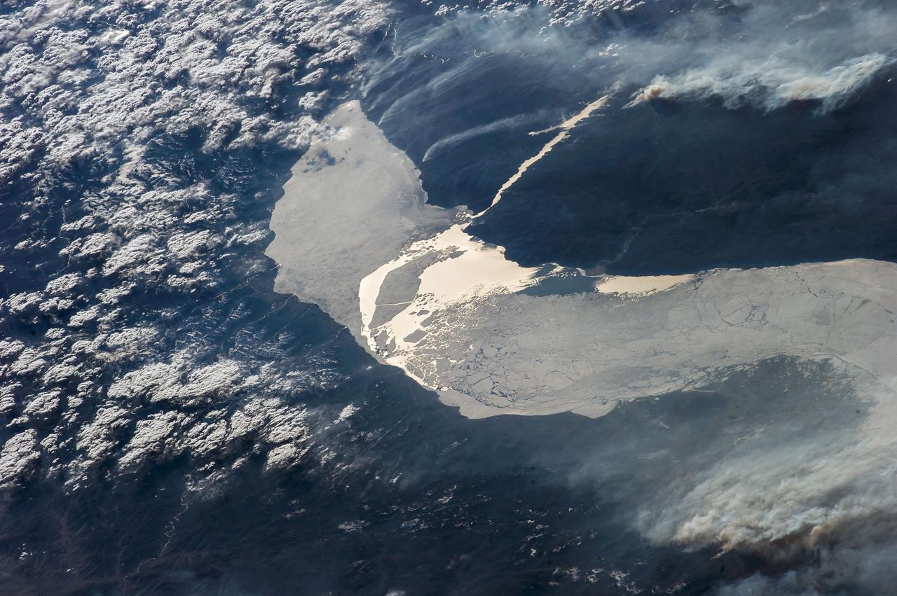 ISS039E14821  (22 April 2014) --- In this late afternoon shot, one of the Expedition 39 crew members aboard the International Space Station looked back toward the setting sun and used a 145mm lens on an electronic still camera to capture the hook-shaped southern half of Lake Baikal in eastern Russia on April 22, 2014.  Most of the lake is covered with dull gray ice.  The brightest point in the center of the image is the reflection of the sun’s rays off a small zone of open water (center) where the ice has begun to melt.  Broken ice masses appear adjacent to the sunglint point (center).  The sun’s rays are also reflected off the water surface of the straight Angara River, the main outlet of the lake (top right). Lake Baikal is 636 kilometers long (395 miles).  It is the deepest lake in the world and holds the largest amount of fresh water. Individual smoke plumes from factories arc away from the north shore (top left). Large smoke palls from wildfires appear in upper right.