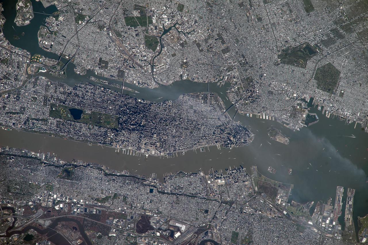 ISS039-E-018541 (5 May 2014) --- New York City is featured in this image photographed by an Expedition 39 crew member on the International Space Station. This detailed image reveals the narrow shape of Manhattan located between the Hudson River and the East River, a feature familiar to crews on the space station. The ragged line of shadow cast by the Palisades cliff crosses the bottom of the image. Wharves jut into the rivers; bridges are visible mainly because of the shadows they cast, and the grid pattern of major roads stands out. On the island of Manhattan itself the main visual features are Central Park (with playing fields as white dots) and two darker zones where the tallest buildings in Midtown East and the Financial District cast strong shadows, even in this early afternoon view. Rivers and parks reduce the effect of the urban heat island – the local zone of higher surface and atmospheric temperatures generated by storage and later release of heat by city materials such as concrete and tarmac. Rivers provide pathways for wind and the cooling effect of parks is detectable by instruments on spacecraft that can measure the temperature of the ground surface. Tall buildings have a more complex effect. Shadowed zones in the "urban canyons" between tall buildings – as shown in this image – receive fewer hours of direct sun per day. But where the sun can reach canyon floors, the sun's energy is reflected back up at the walls of the buildings where it is absorbed and later released as heat. This is especially the case at night when urban canyons retain more heat than those sections of the city with shorter buildings.