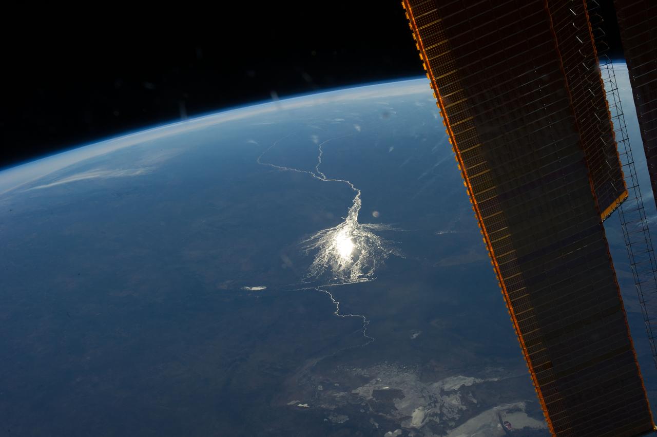 ISS040-E-008209 (6 June 2014) --- Okavango inland delta in northern Botswana is featured in this image photographed by an Expedition 40 crew member on the International Space Station. The great Okavango delta in the Kalahari Desert is illuminated in the sun?s reflection point in this panorama. Using this sun glint technique, crew members can image fine detail of water bodies. Here the bright line of the Okavango River shows the annual summer flood advancing from the well-watered Angolan Highlands (upper margin) to the delta. Then the flood water slowly seeps across the 150 kilometer-long delta, supplying forests and wetlands, finally reaching the fault-bounded lower margin of the delta in the middle of winter. Most of the water of this large river is used up by the forests, or evaporates in the dry air. Only two percent of the river?s water actually exits the delta. The wetland supports high biodiversity in the middle of the otherwise semiarid Kalahari Desert, and is now one of the most famous tourist sites in Africa. This view also shows the small quantity of water in the Boteti River. Okavango water only reaches the dry lake floors (lower right) in the wettest years. Part of one of the station?s solar arrays is visible at right.